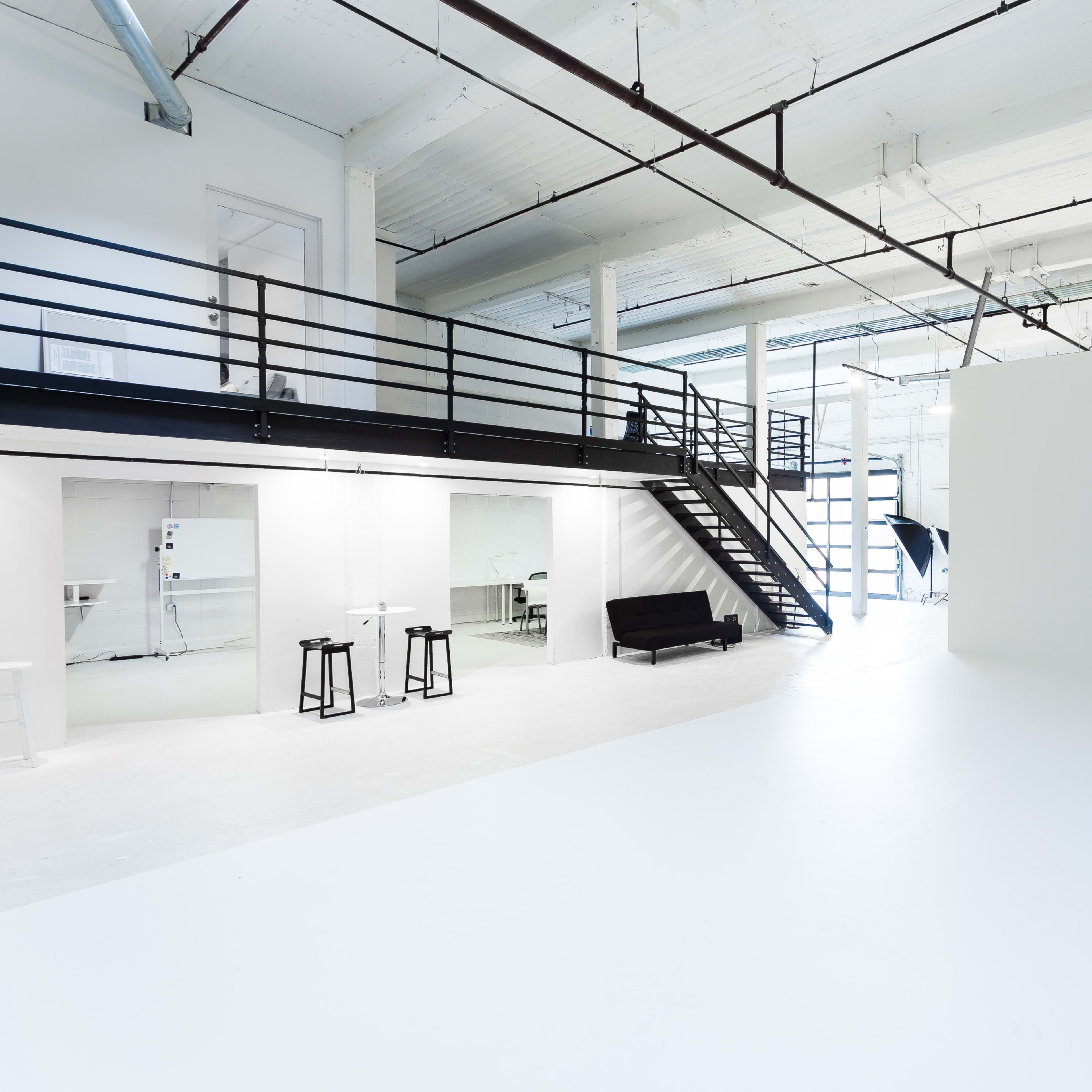 A spacious, modern studio with white walls, a staircase leading to a mezzanine, and minimalist furniture.