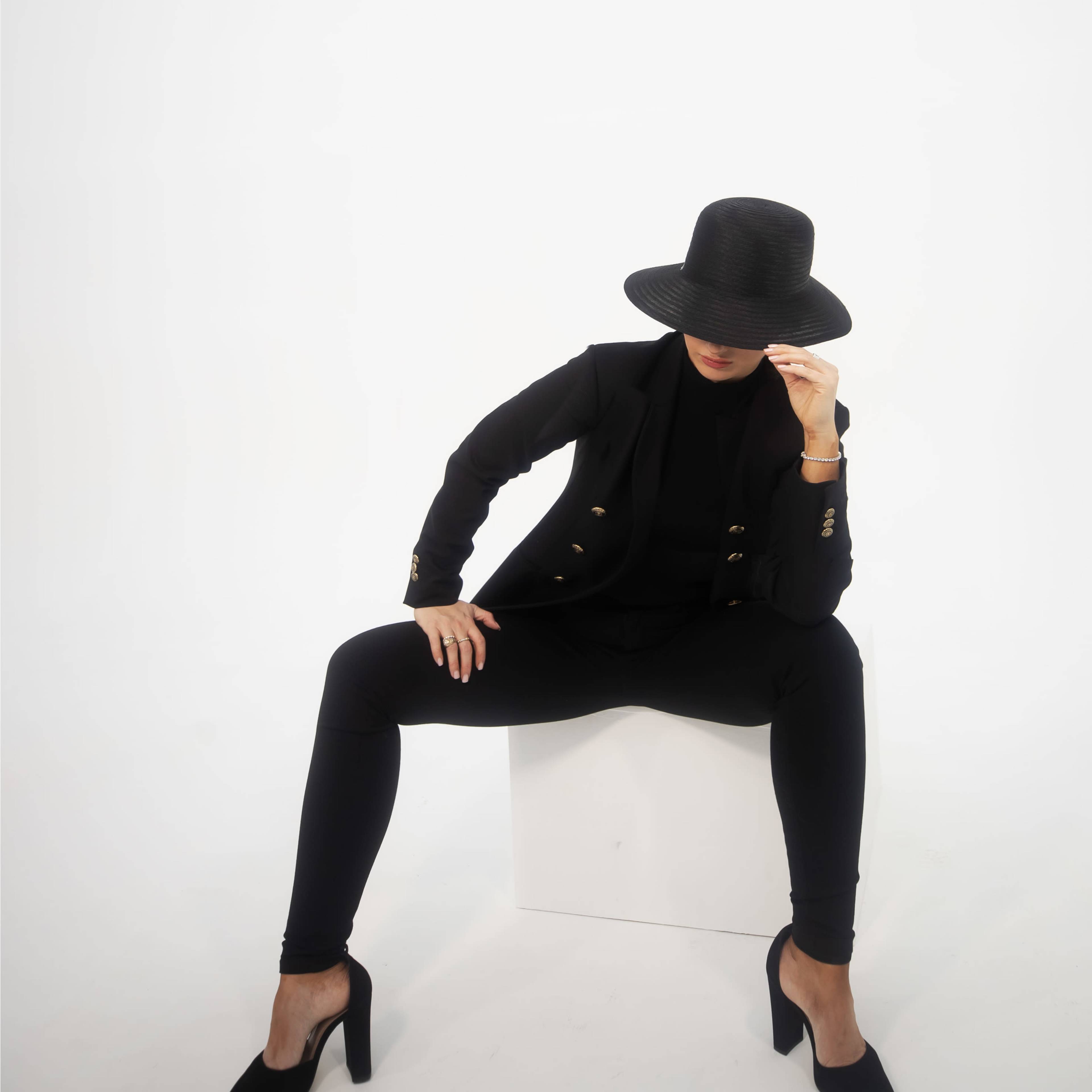 A woman in a black suit and hat sits on a white cube, with her legs crossed and one hand adjusting her hat.