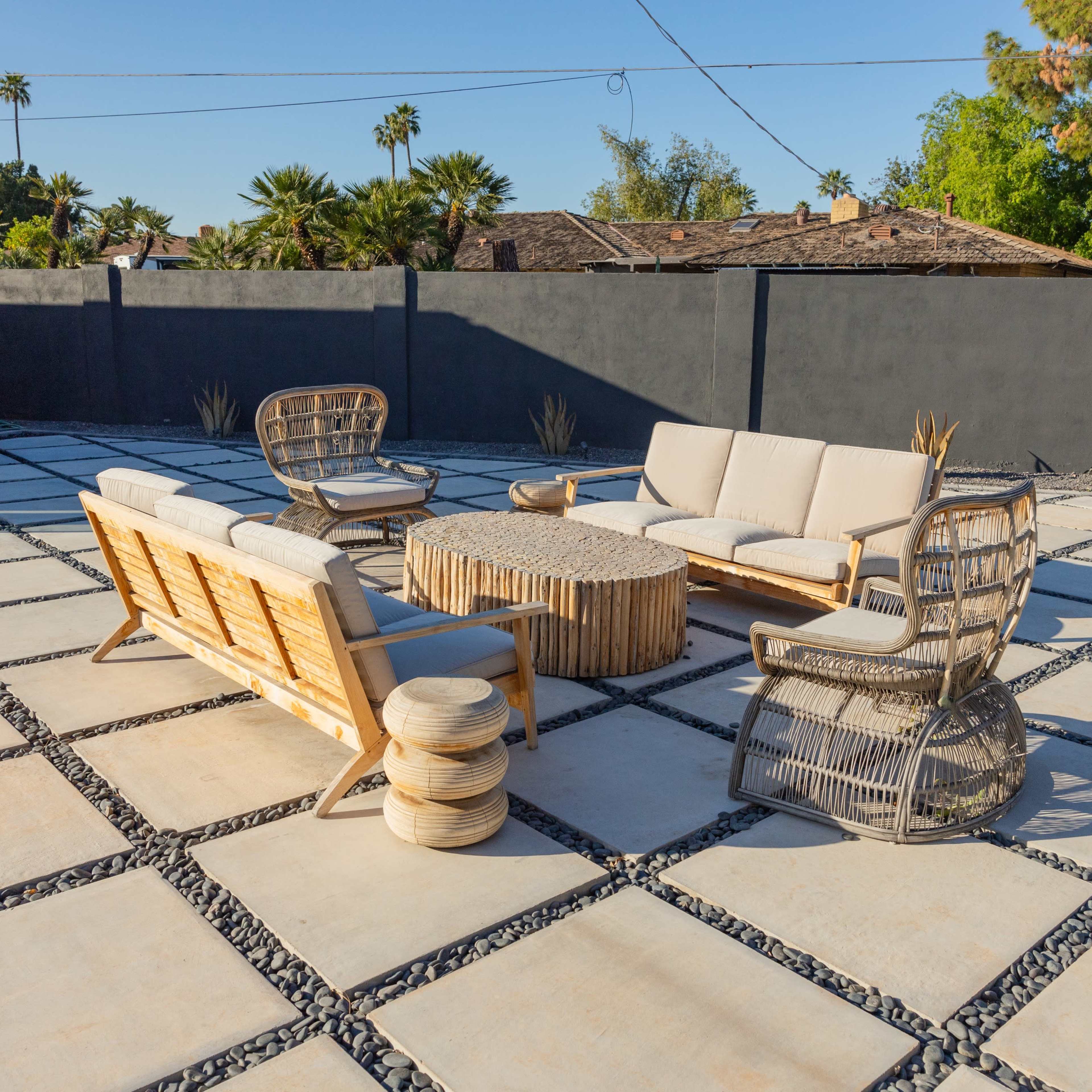 A patio area features wooden and wicker furniture arranged around a circular table, set on a patterned stone surface with gravel in between.
