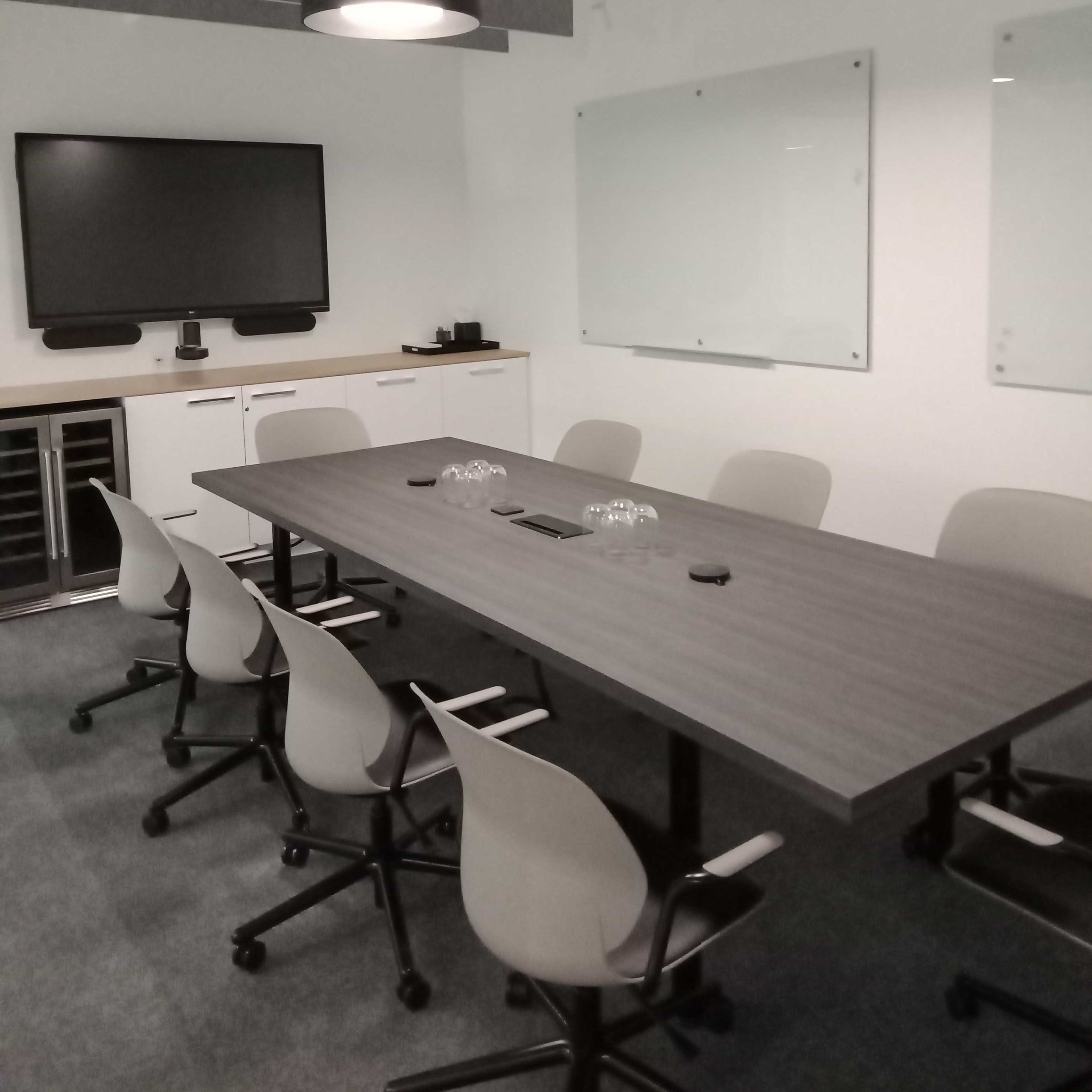 A modern conference room features a large table surrounded by eight chairs, with a television and whiteboards on the walls.