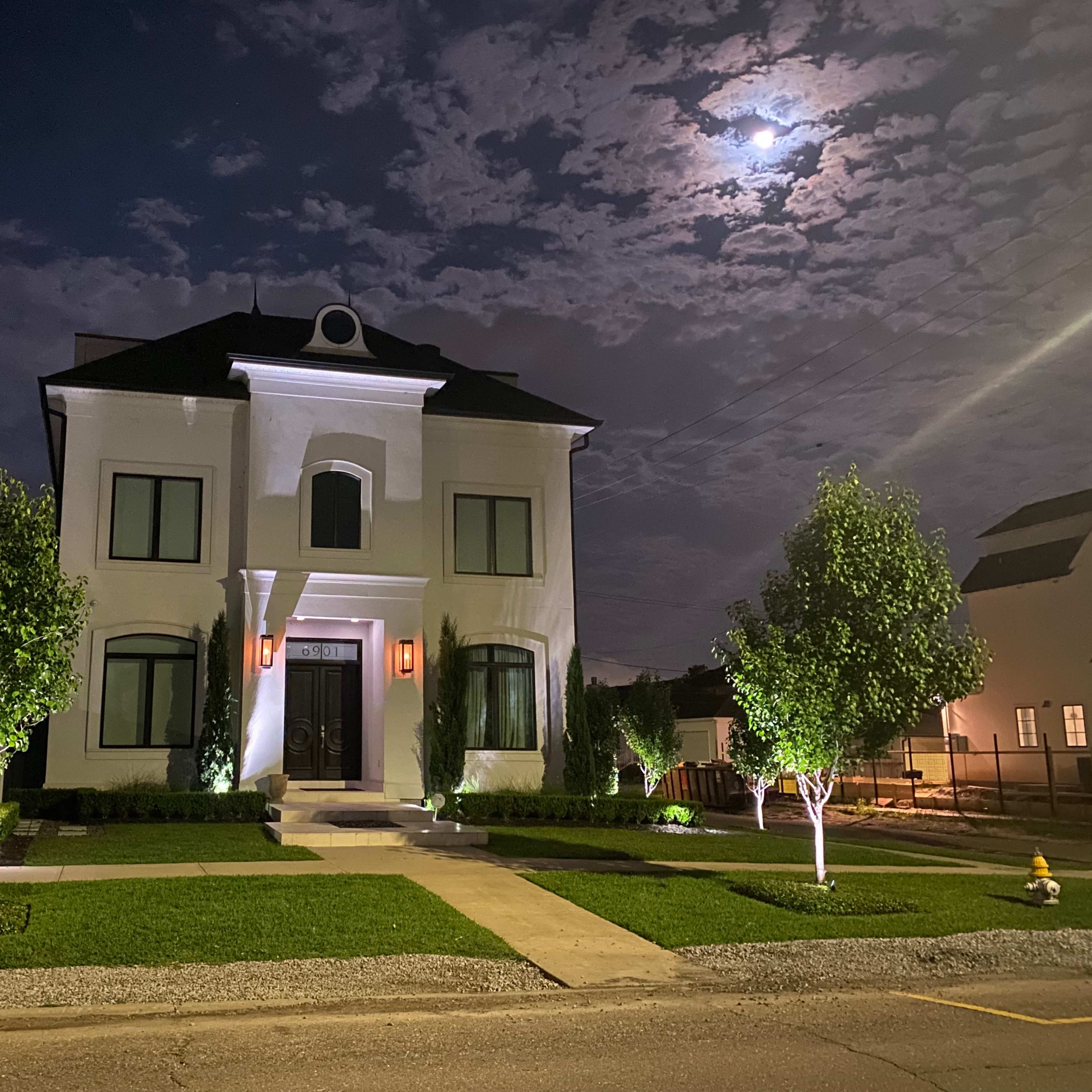 A well-lit, two-story white house stands at night under a partly cloudy sky, with two small trees illuminated on either side of the pathway leading to the front door.