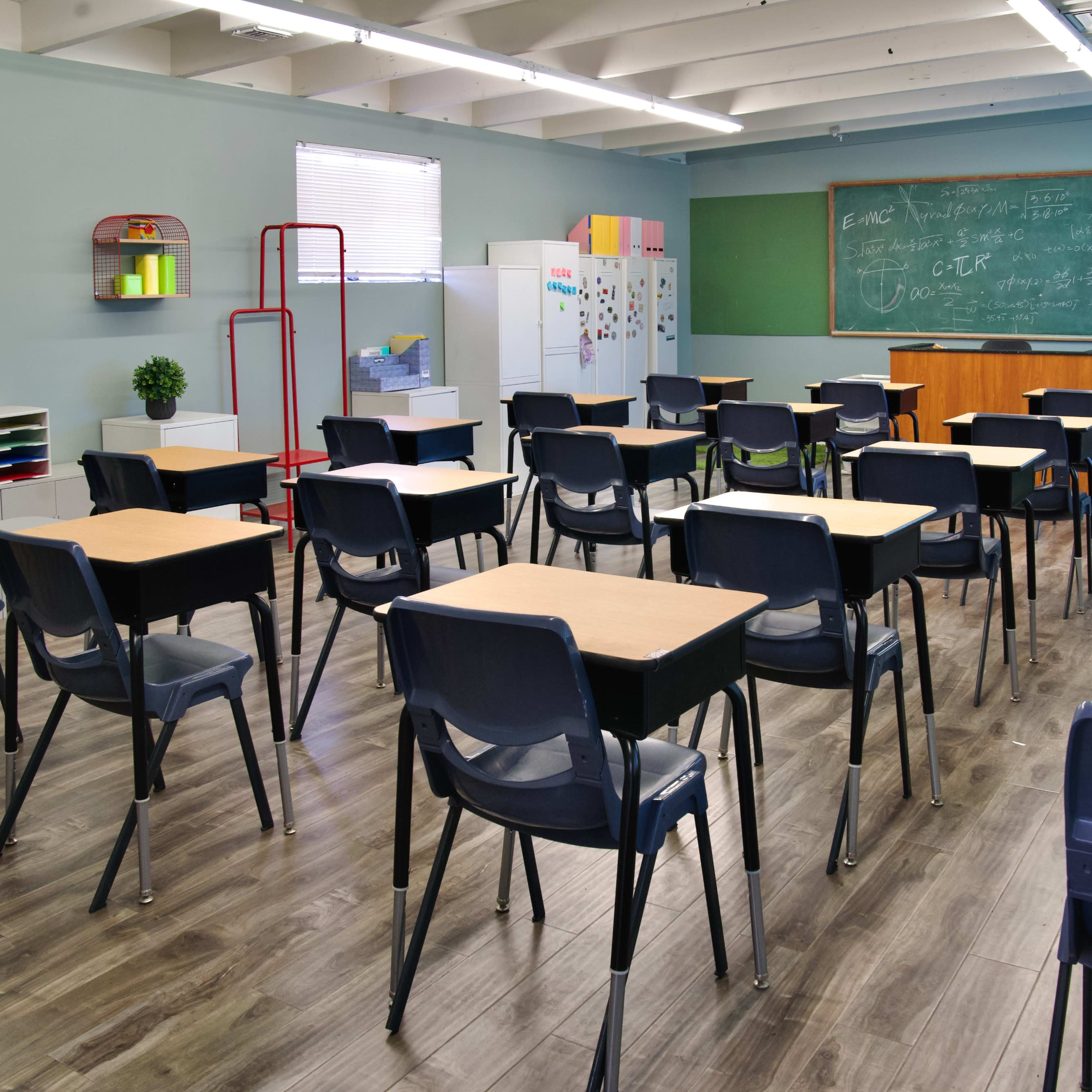 A classroom with empty black chairs and wooden desks arranged in rows, a chalkboard at the front, and various educational materials along the walls.
