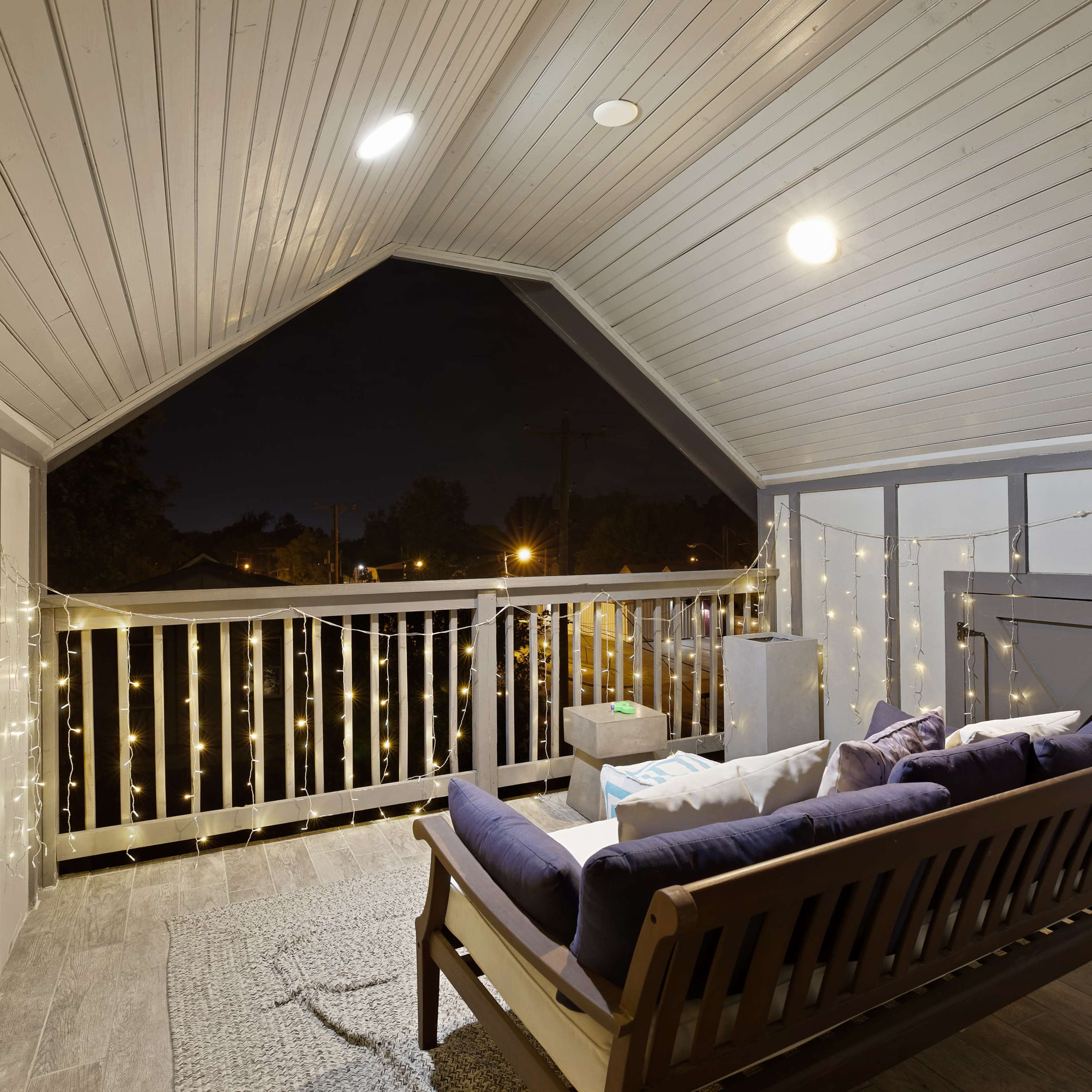 A cozy balcony features a wooden bench, string lights, and a view of the night sky.
