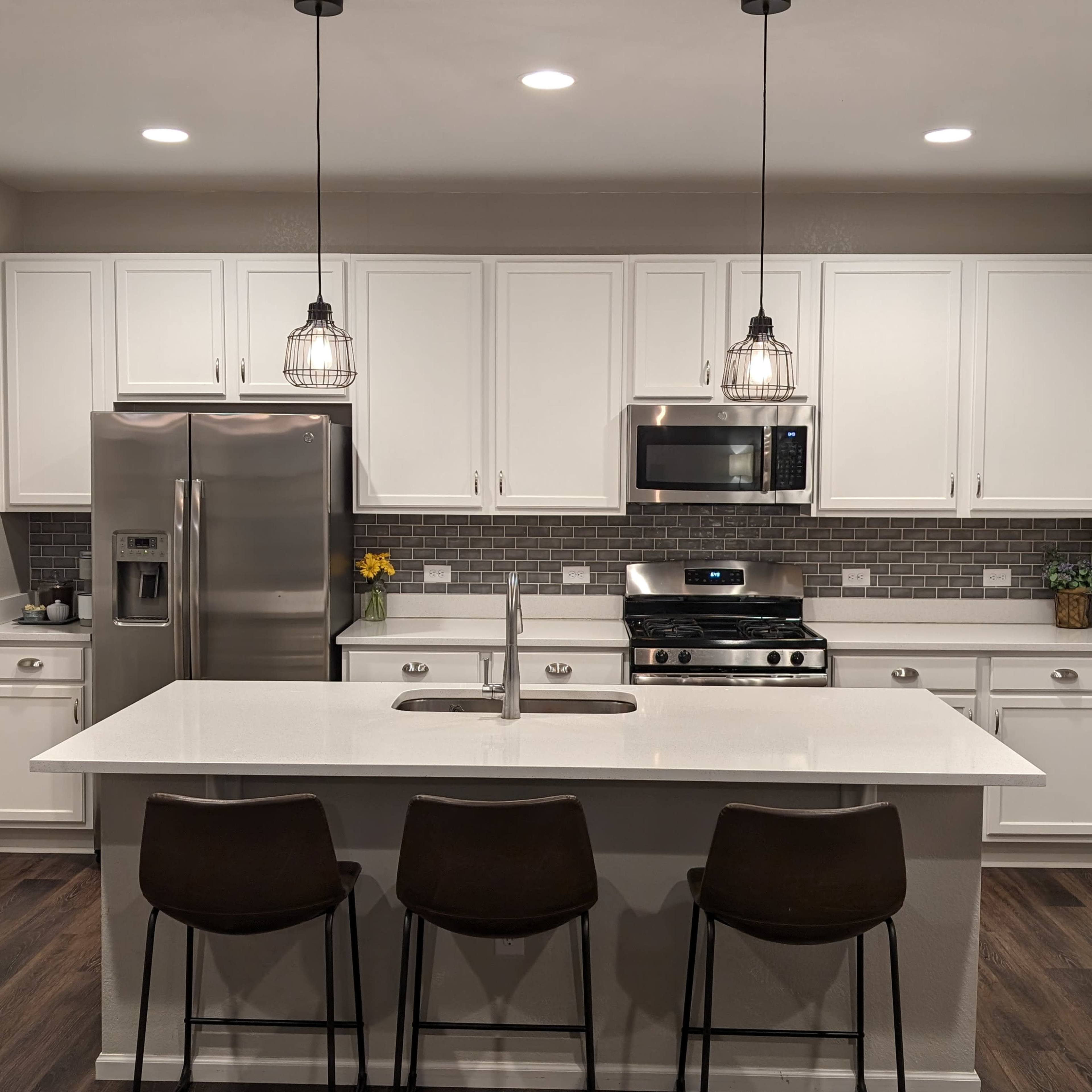 A modern kitchen featuring white cabinets, stainless steel appliances, a central island with three bar stools, and pendant lighting.