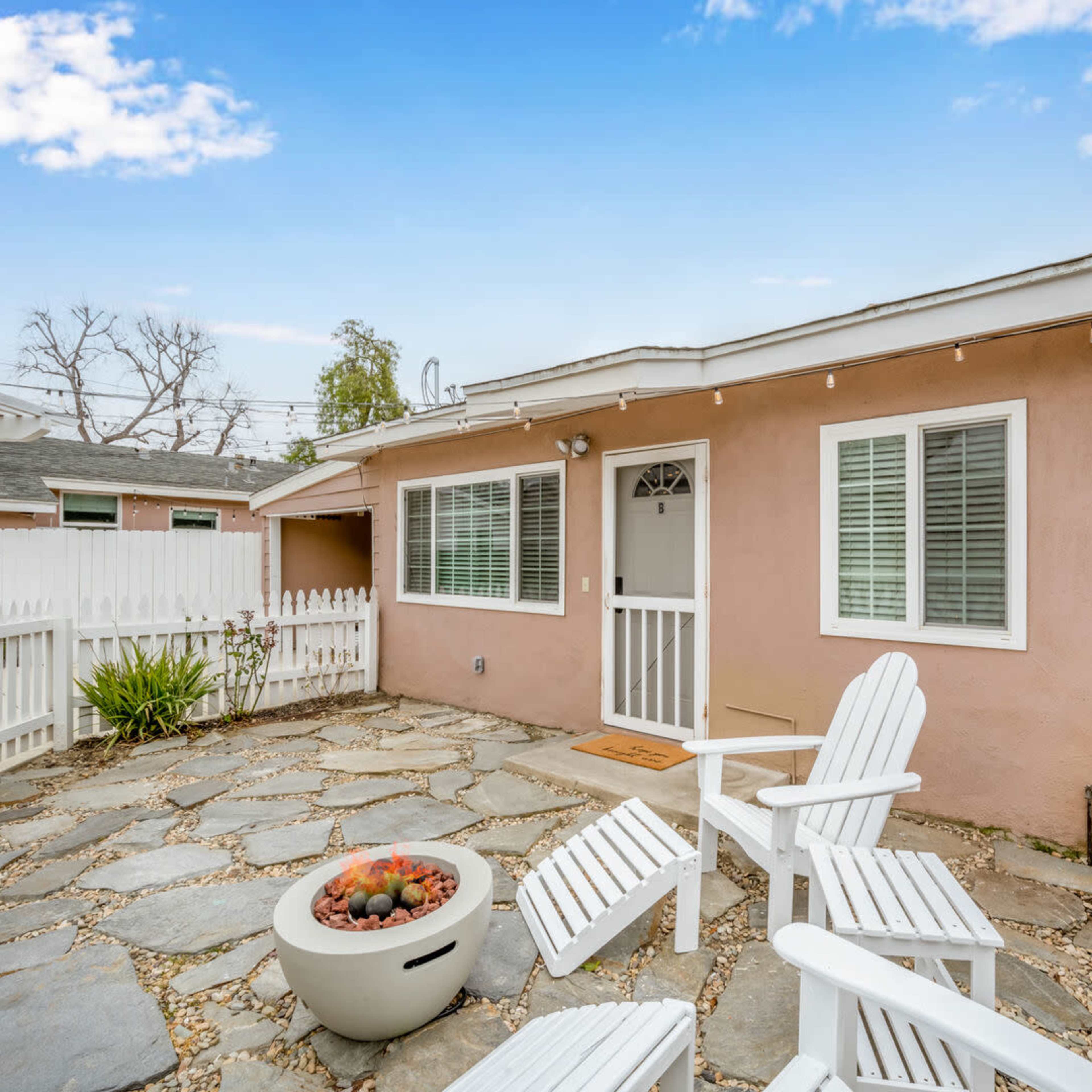 A stone patio with two white adirondack chairs and a fire pit is situated beside a peach-colored house with several windows.
