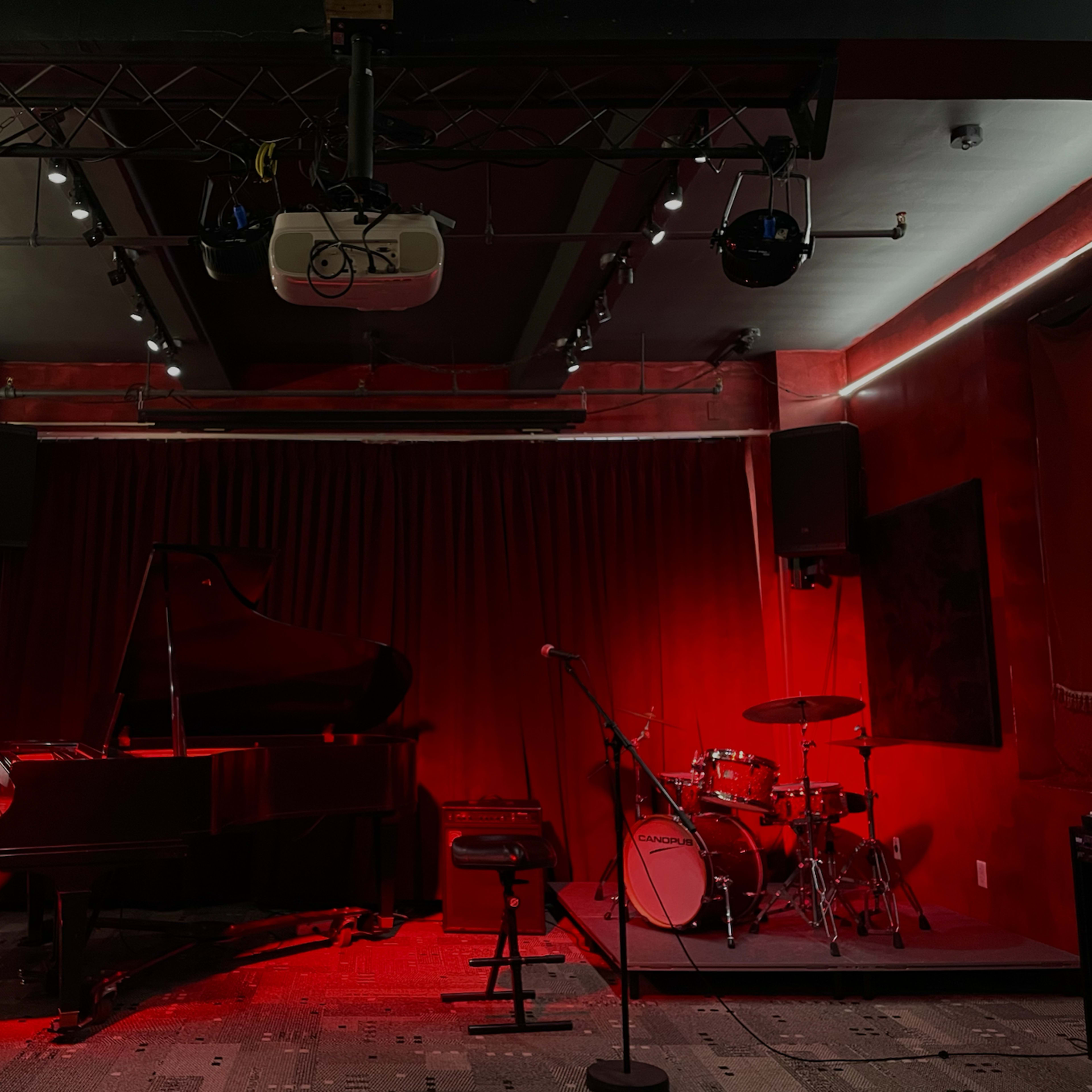 A dimly lit music venue features a grand piano, a drum set, and red walls with sound equipment mounted above.
