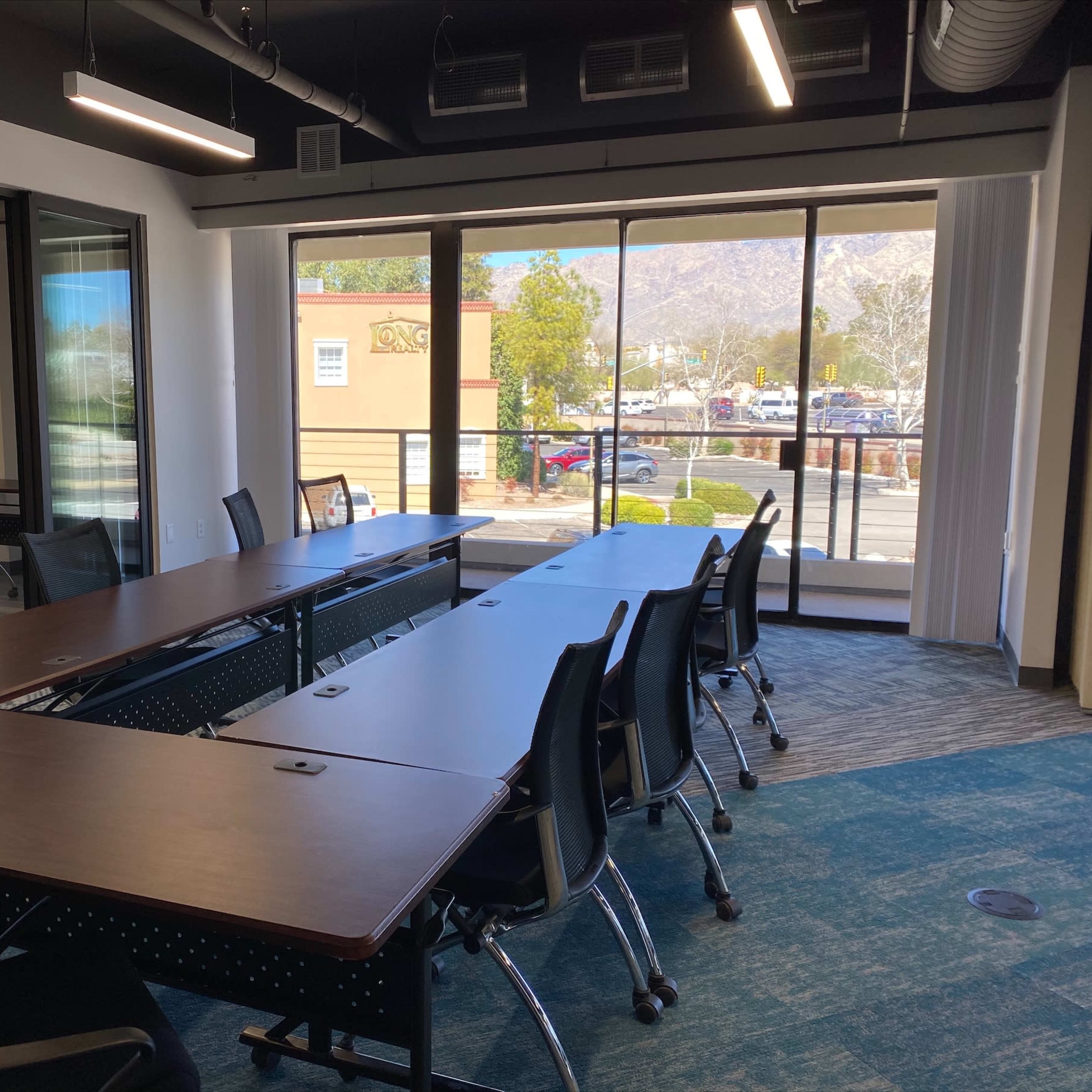 A conference room with several tables arranged in a U-shape and a large window that offers a view of a parking lot and mountains in the background.