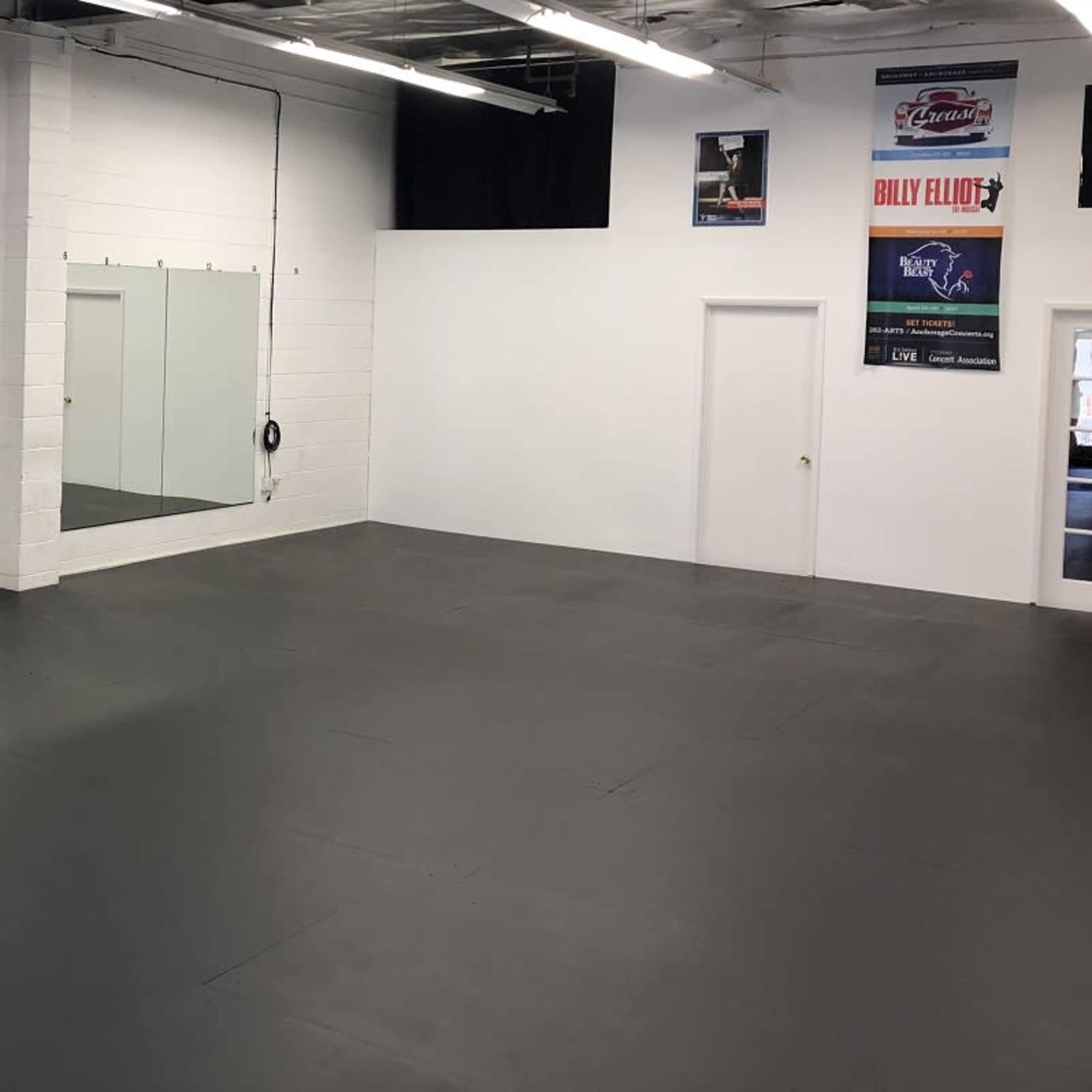 An empty, well-lit room with gray flooring, large mirrors on one wall, and two doors visible in the background.