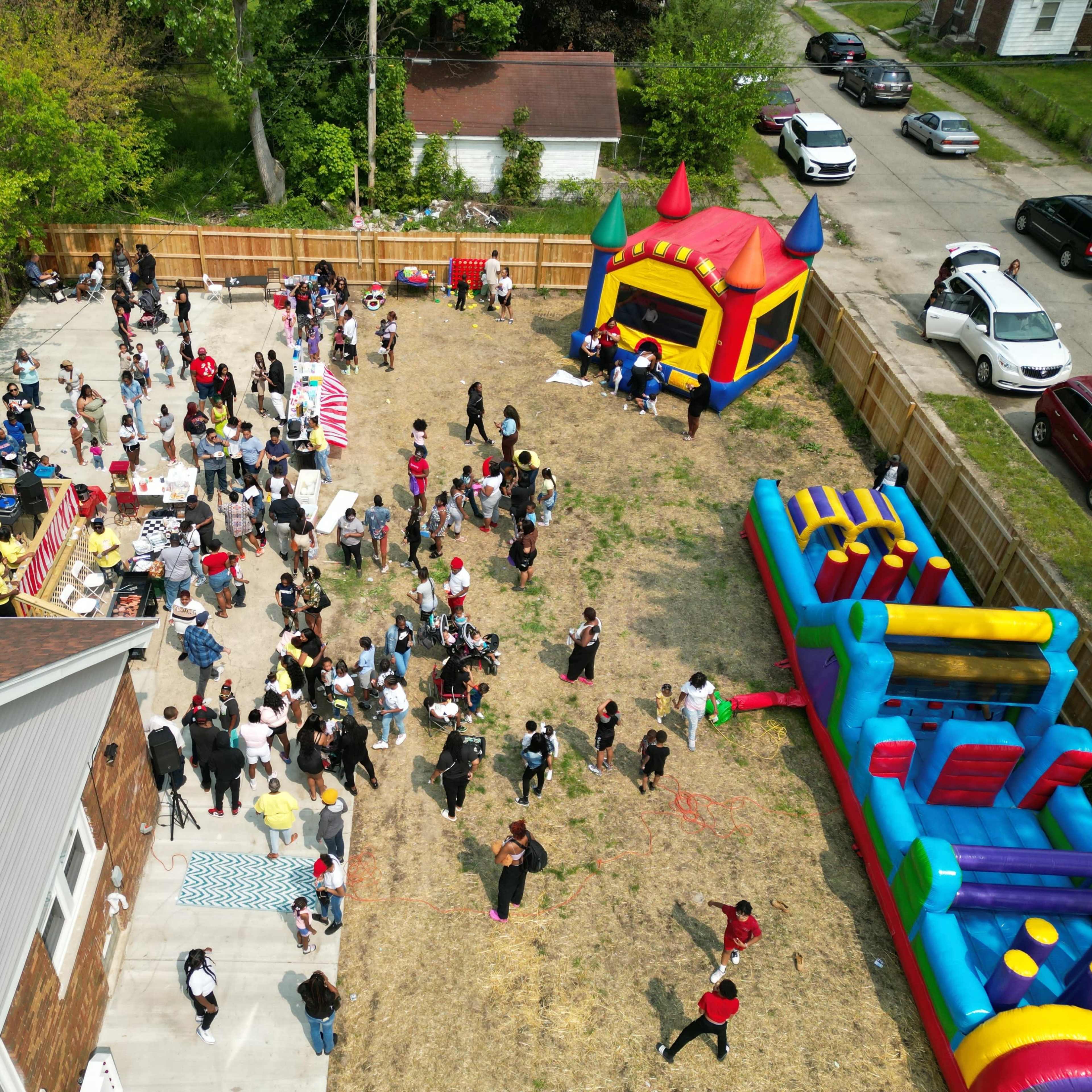 A large group of people gathers in a backyard filled with a bounce house, food stalls, and festive activities.