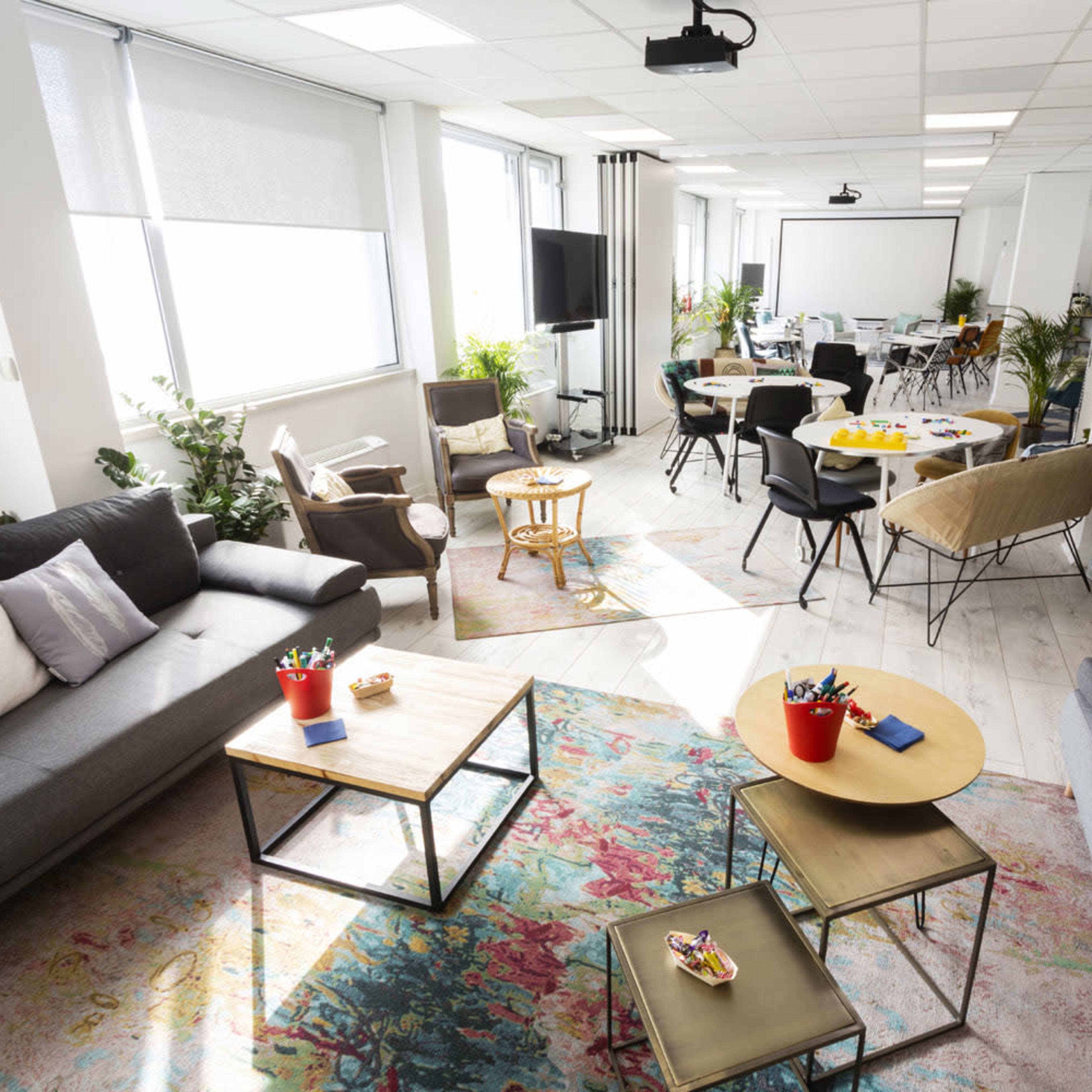 A bright and modern workspace features comfortable seating areas, tables for meetings, and plants throughout the room.