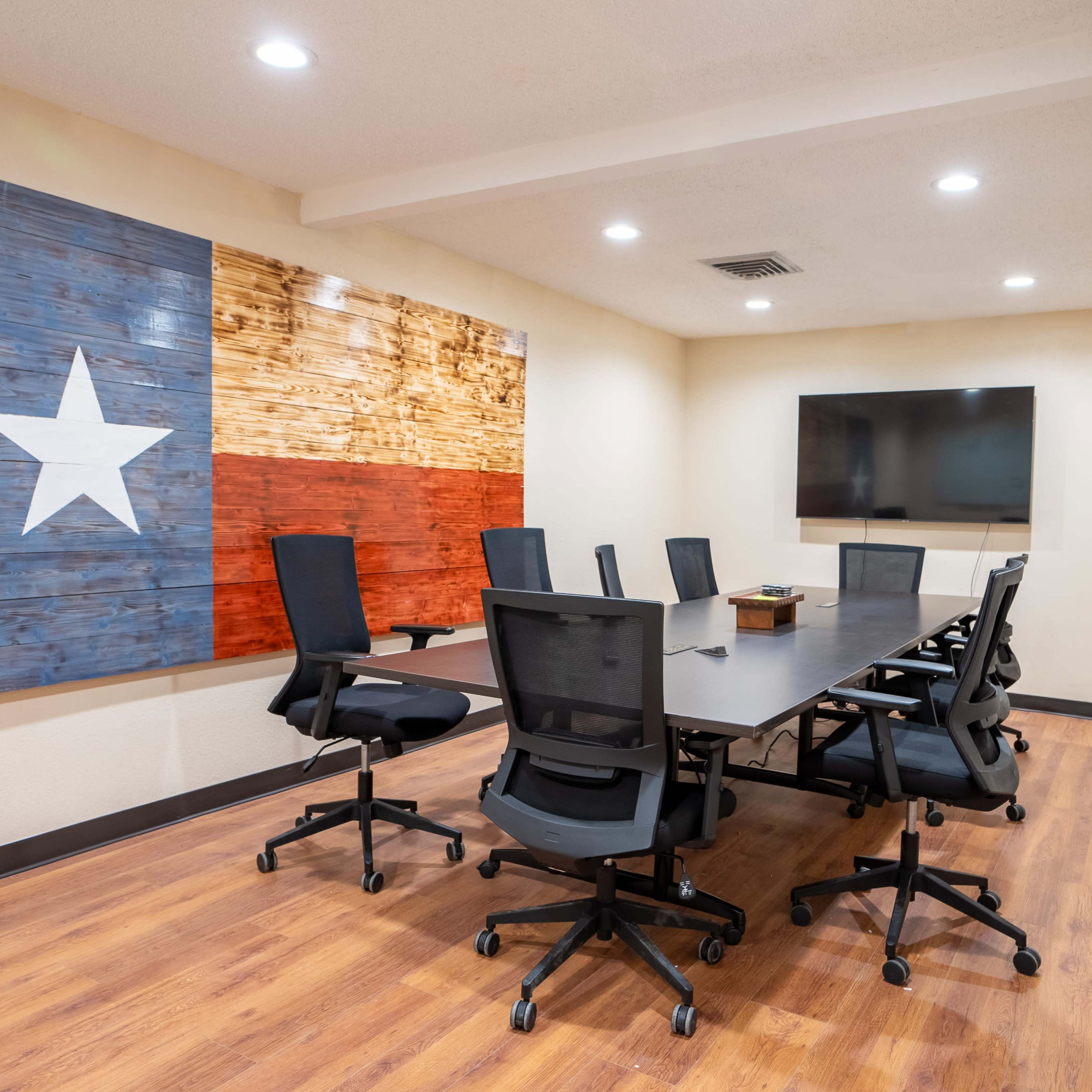A conference room features a large table surrounded by black chairs, with a Texas flag mural on the wall and a wall-mounted monitor.