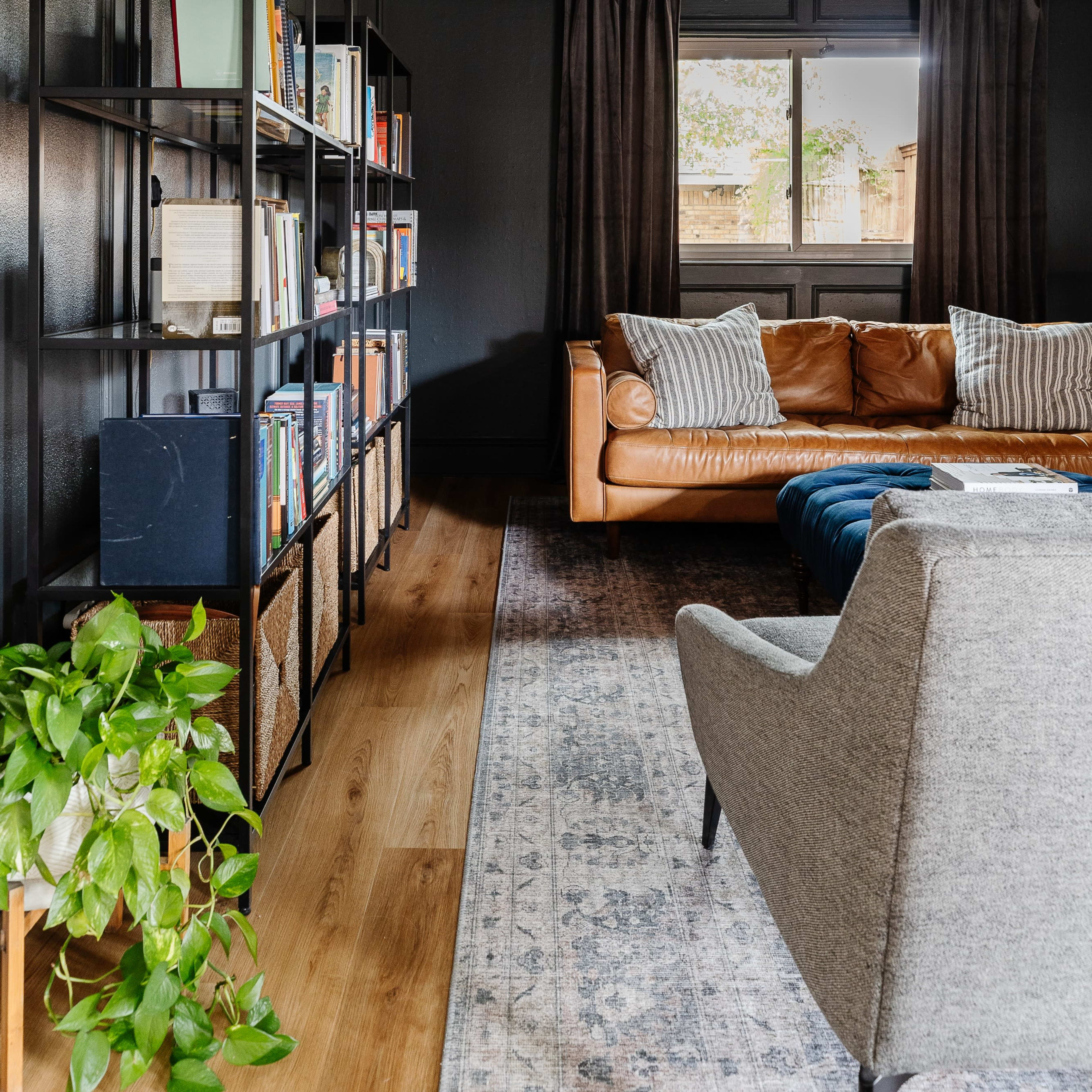A modern living room features a leather sofa, a gray armchair, a tall bookshelf with books, and a large window with dark curtains.