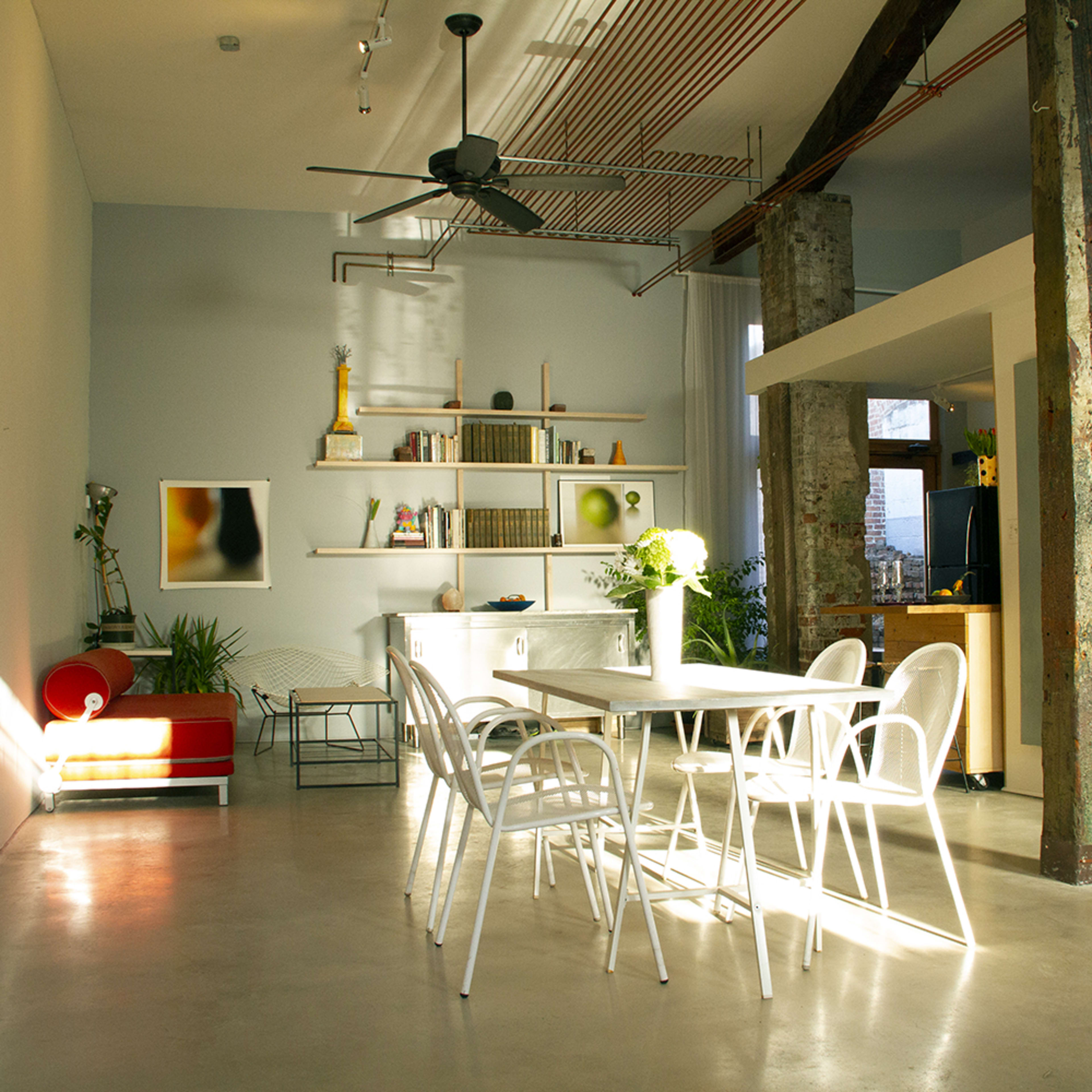 A modern, minimalist dining area with a large table surrounded by white chairs, a kitchenette in the background, and natural light streaming through the windows.