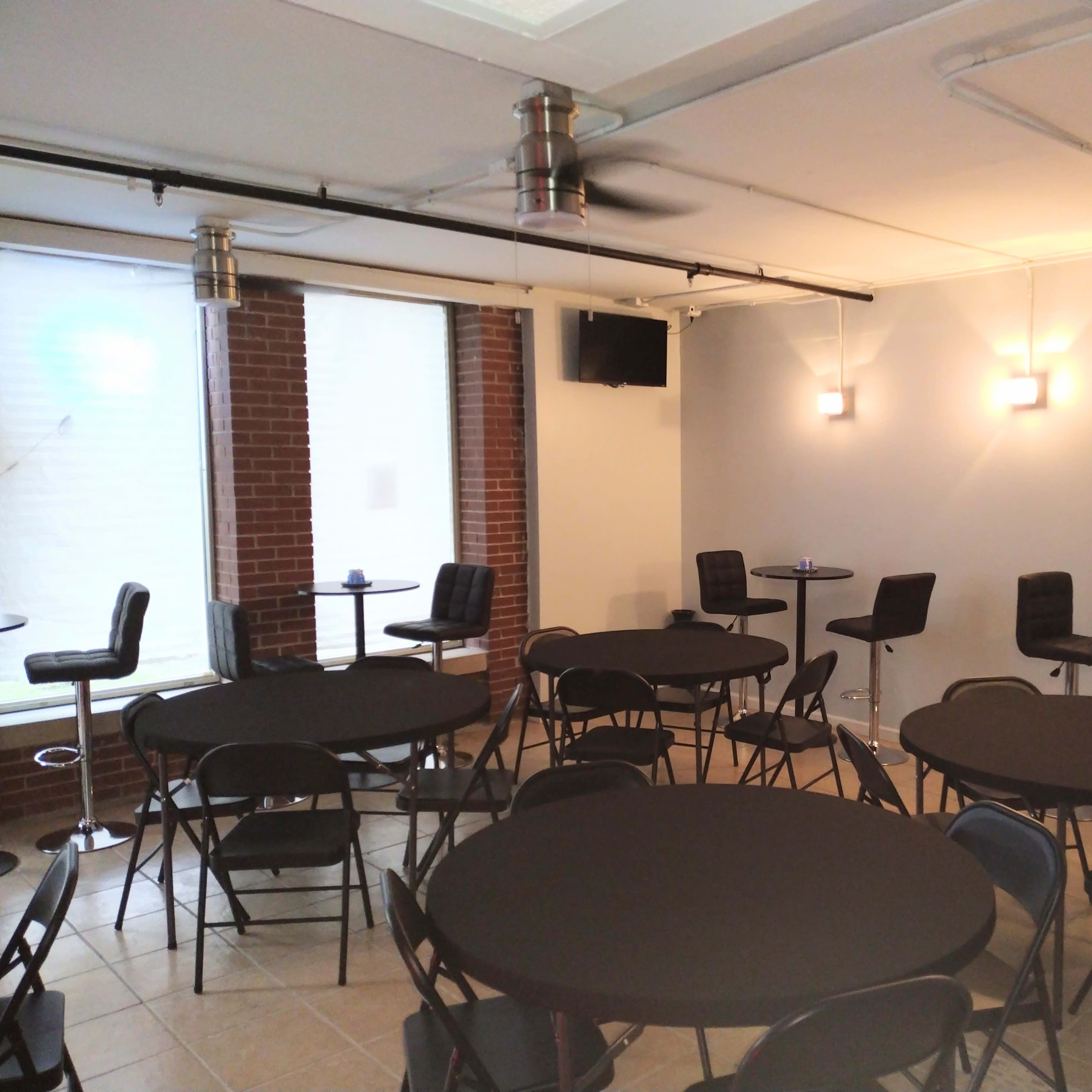 A spacious room with several circular tables covered in black tablecloths and high-top chairs arranged around them, illuminated by wall-mounted lights.