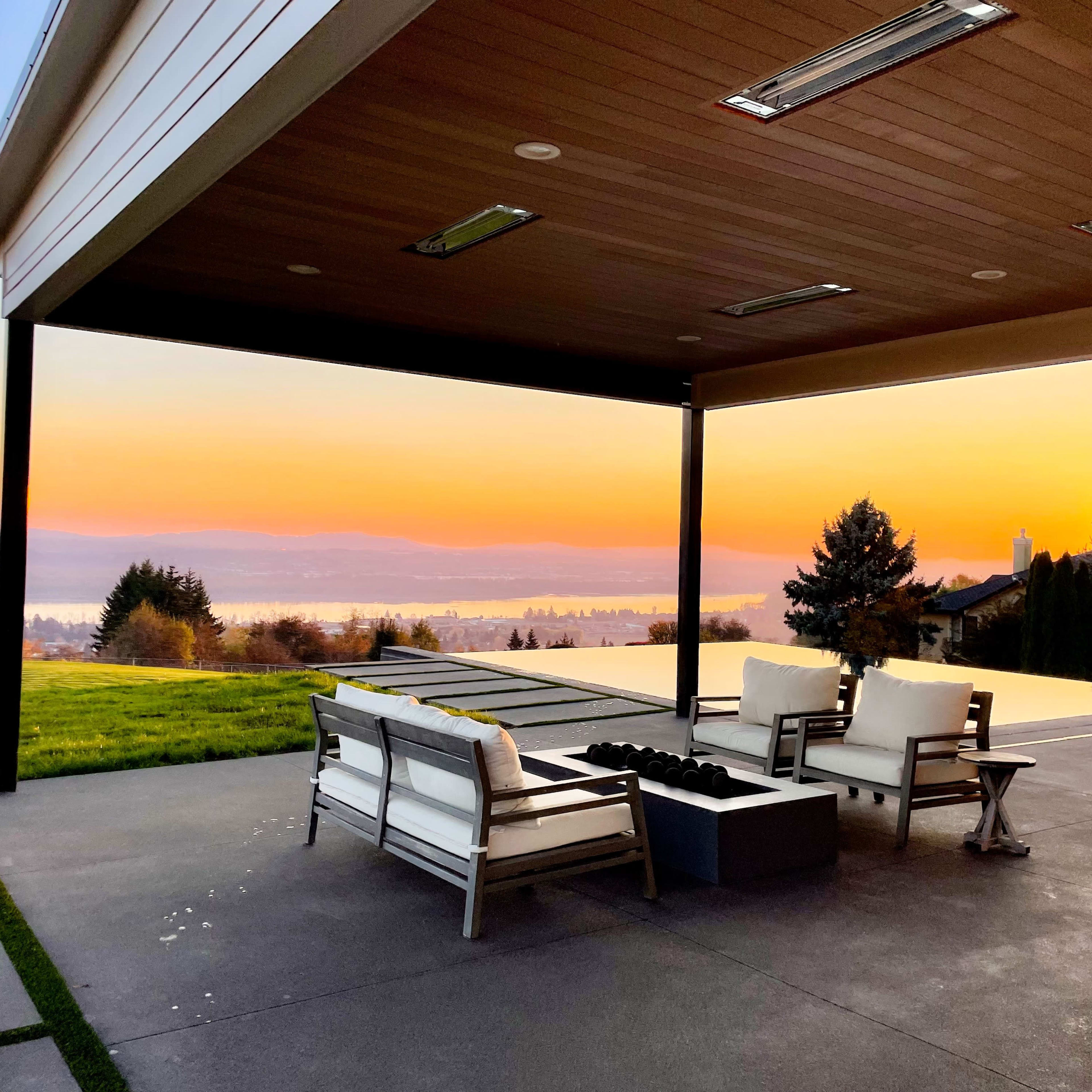 A covered patio with white furniture and a fire pit overlooks a sunset view of a lake and distant hills.