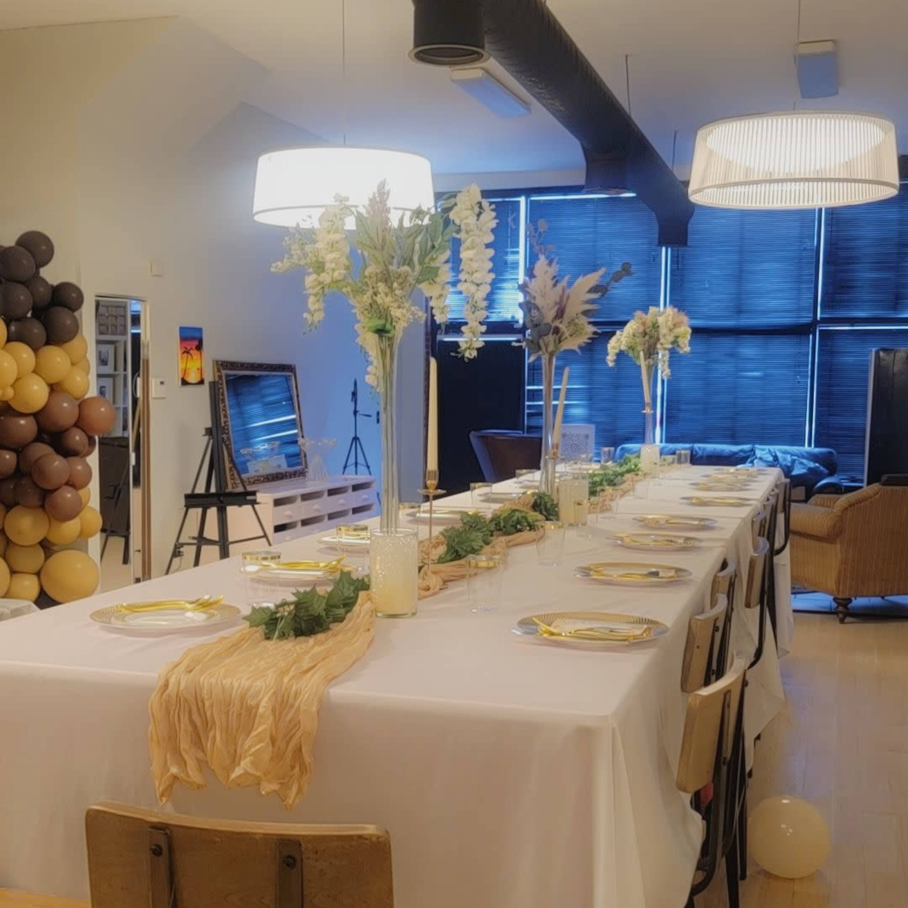 A long table is set for a gathering, adorned with a white tablecloth, candles, and floral arrangements, in a well-lit room featuring decorative balloons in the background.
