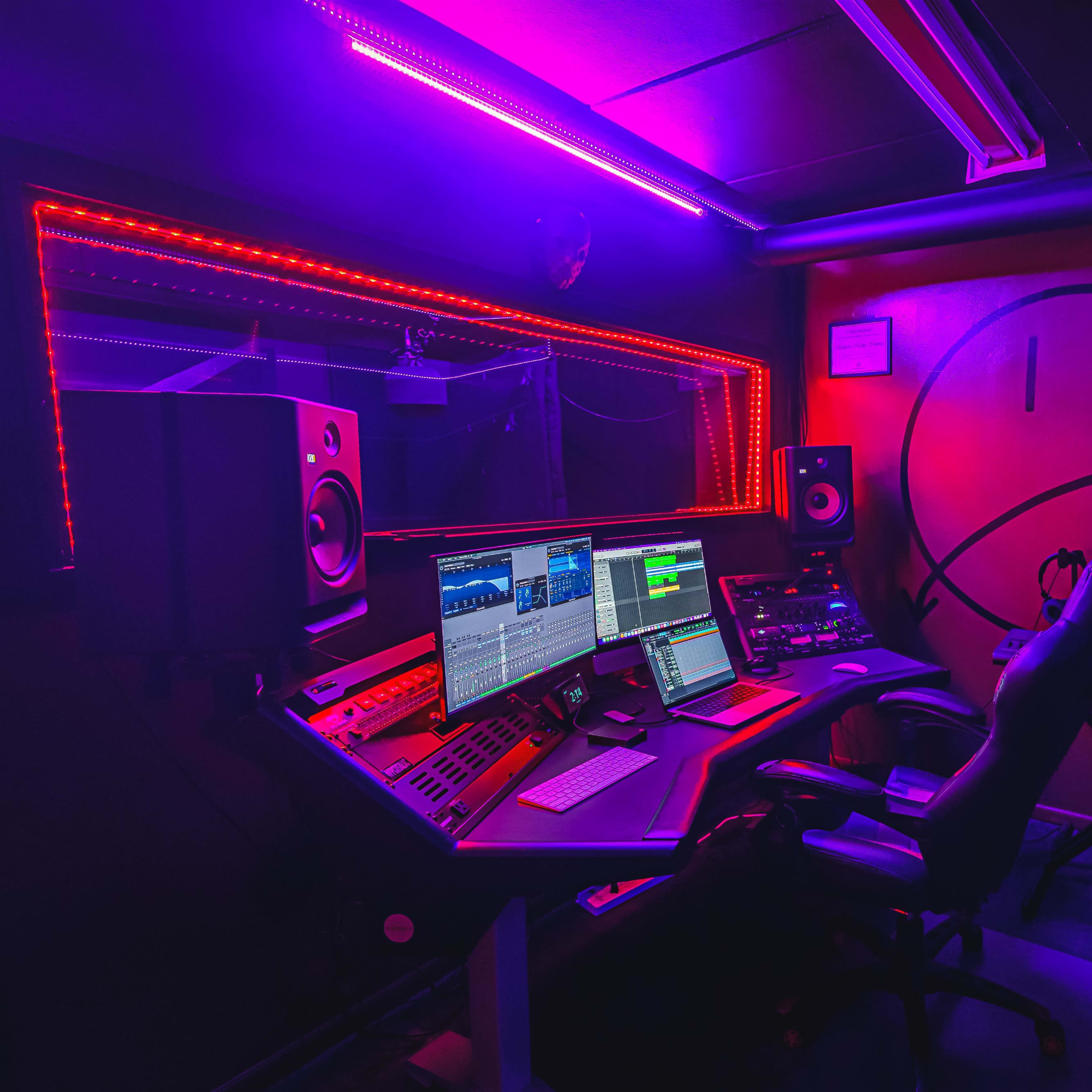A dimly lit music studio featuring a control console, computer monitors, and colorful LED lights illuminating the room.