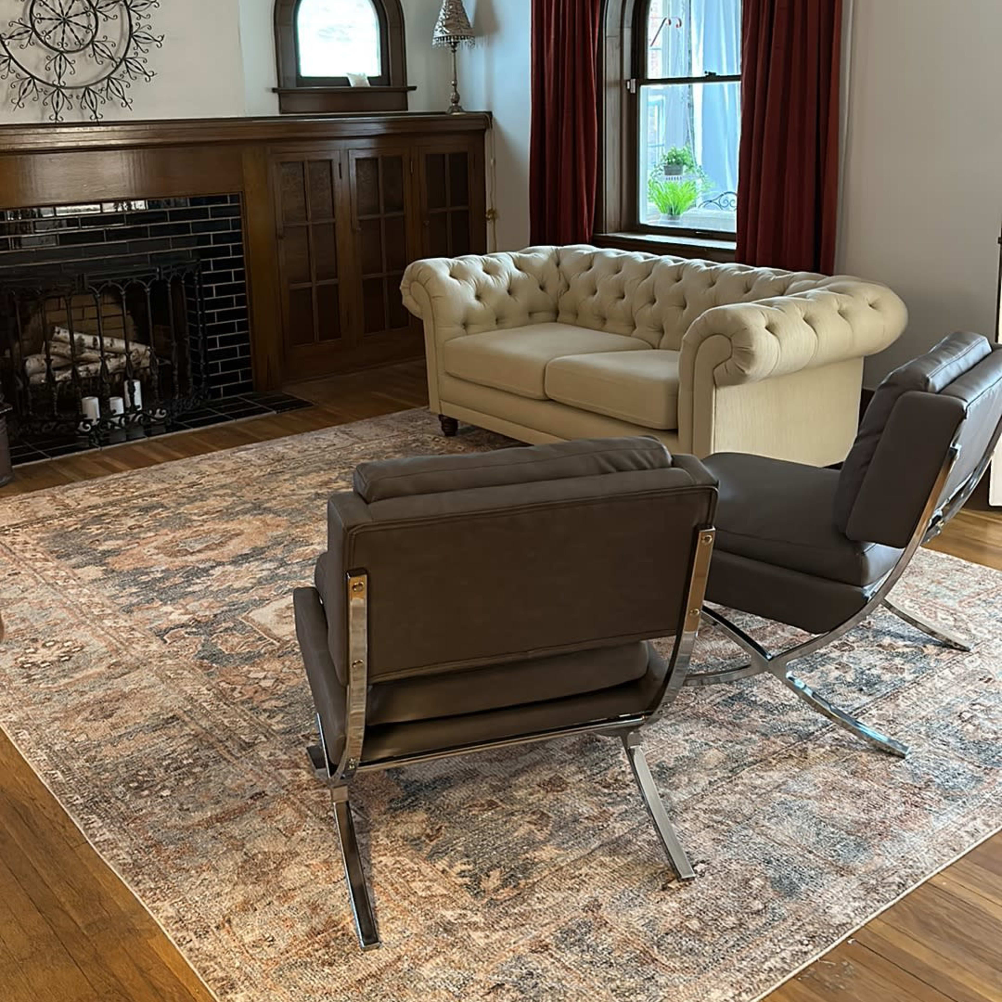 A light-colored tufted sofa sits in a living room with hardwood floors, accompanied by two modern chairs and a large area rug.
