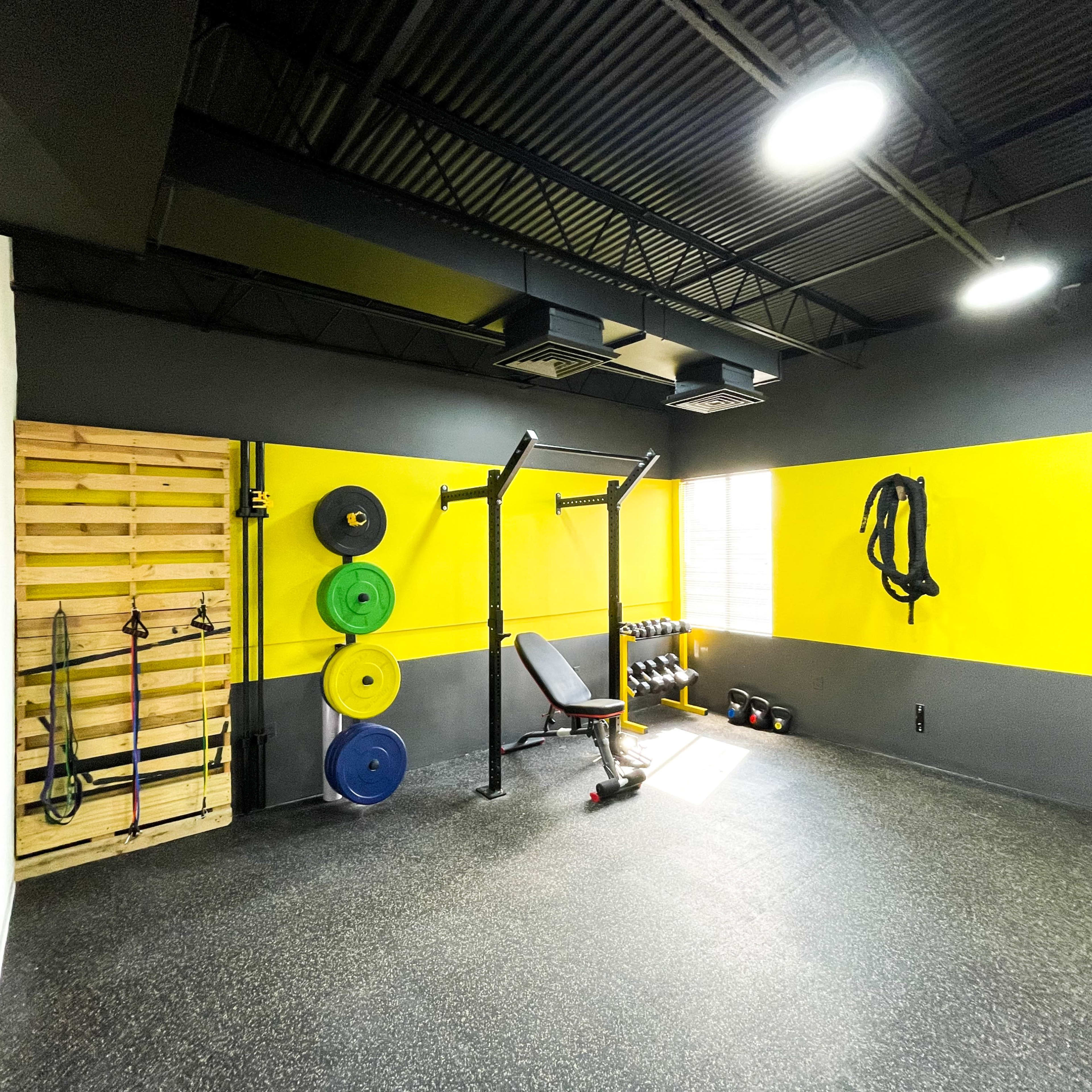 A gym interior with weightlifting equipment, including a squat rack, dumbbells, and resistance bands against a yellow and gray wall.