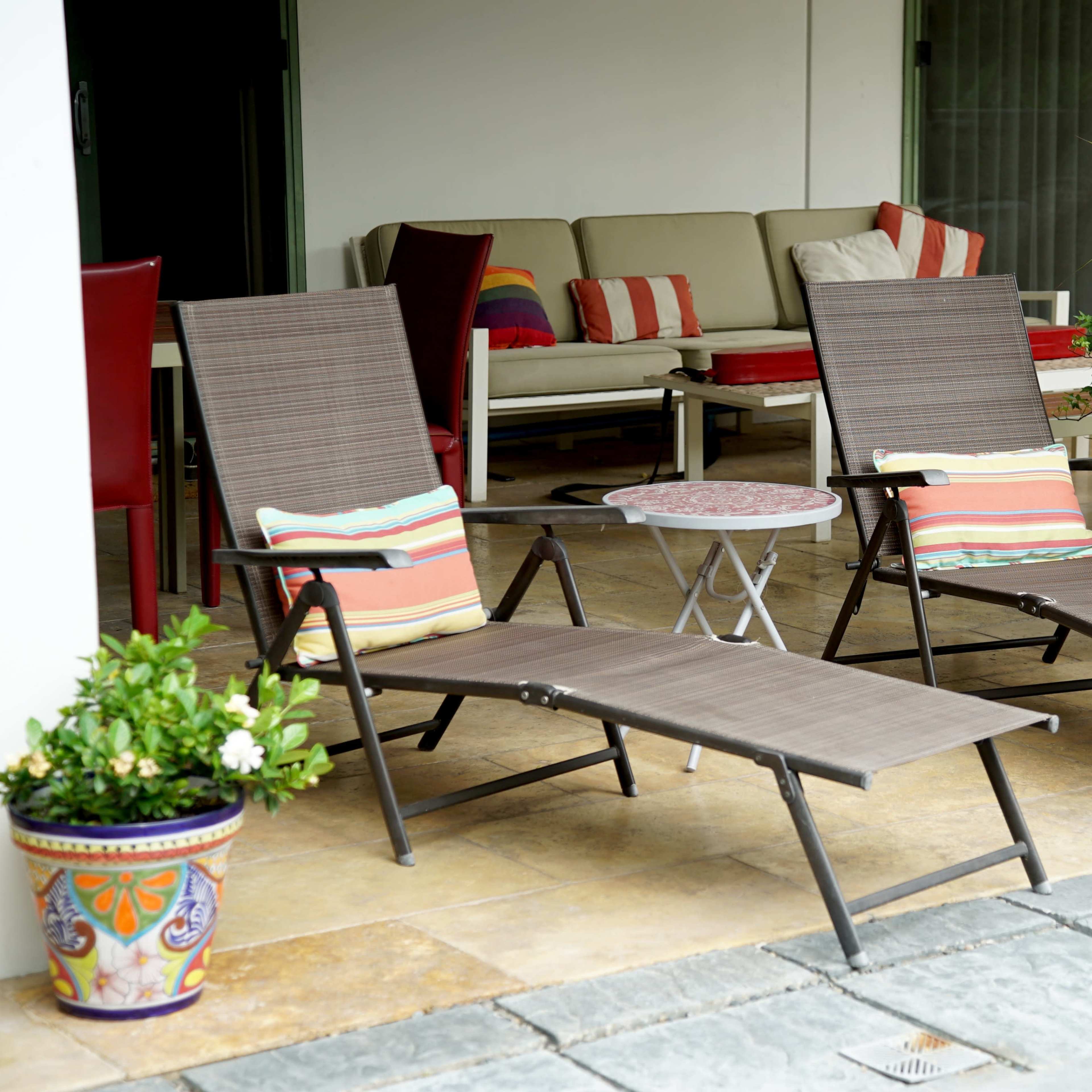 Two reclining lounge chairs with cushions sit beside a small table on a patio covered by a roof, adjacent to a sofa and decorative plants.