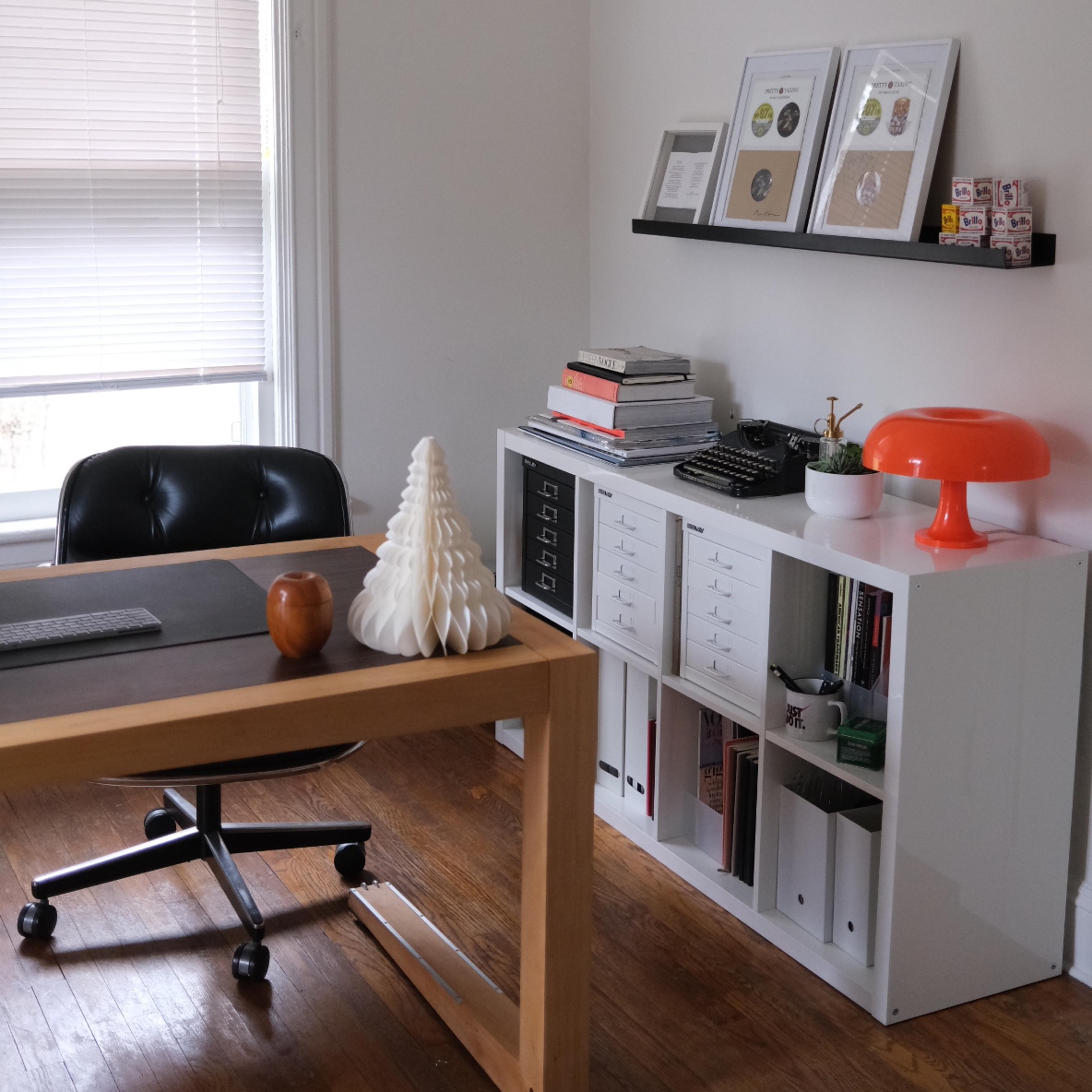 A minimalistic office space featuring a wooden desk with a black chair, a white storage unit, decorative items, and natural light coming through a window.