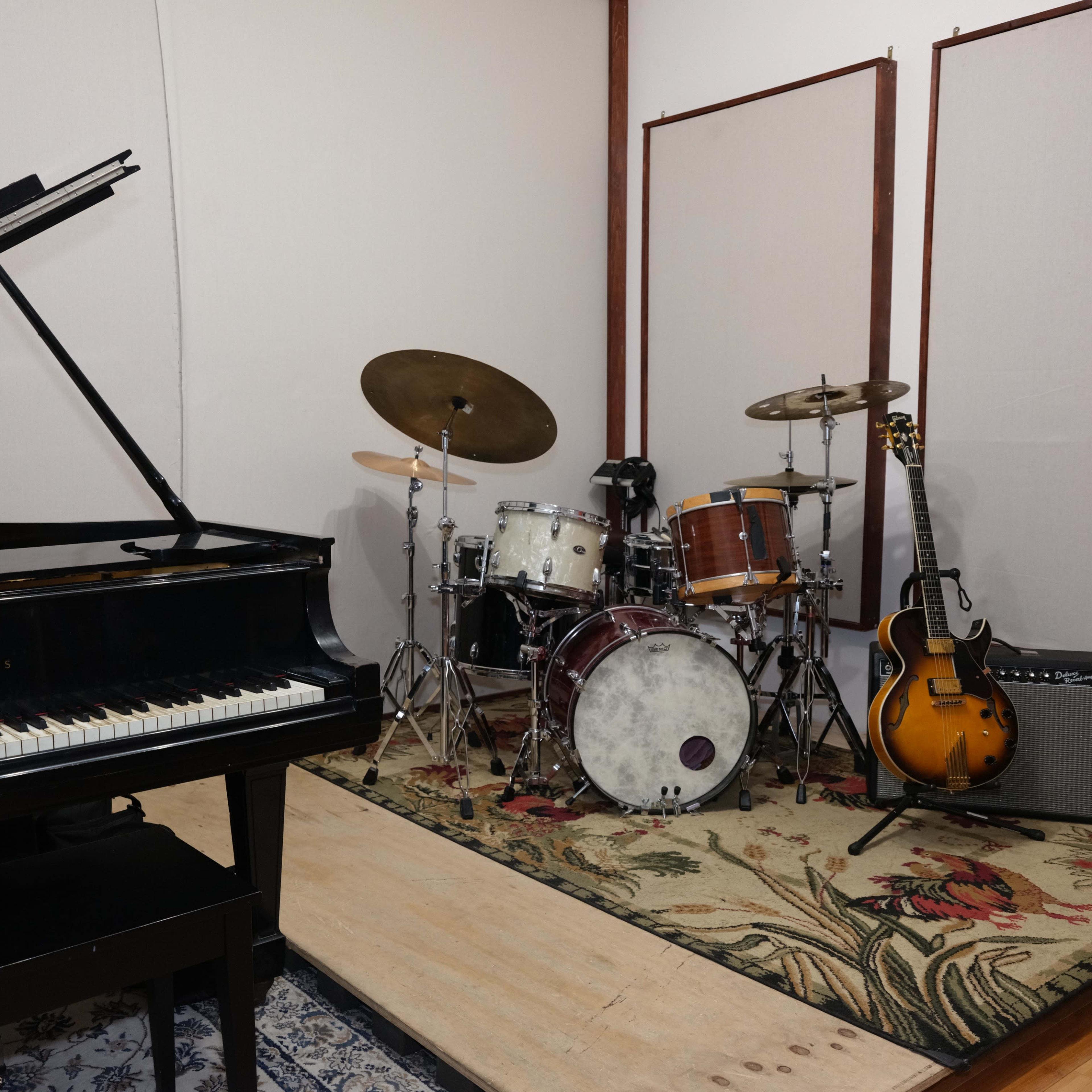 A grand piano is positioned next to a drum set, an electric guitar, and an amplifier in a music rehearsal space.