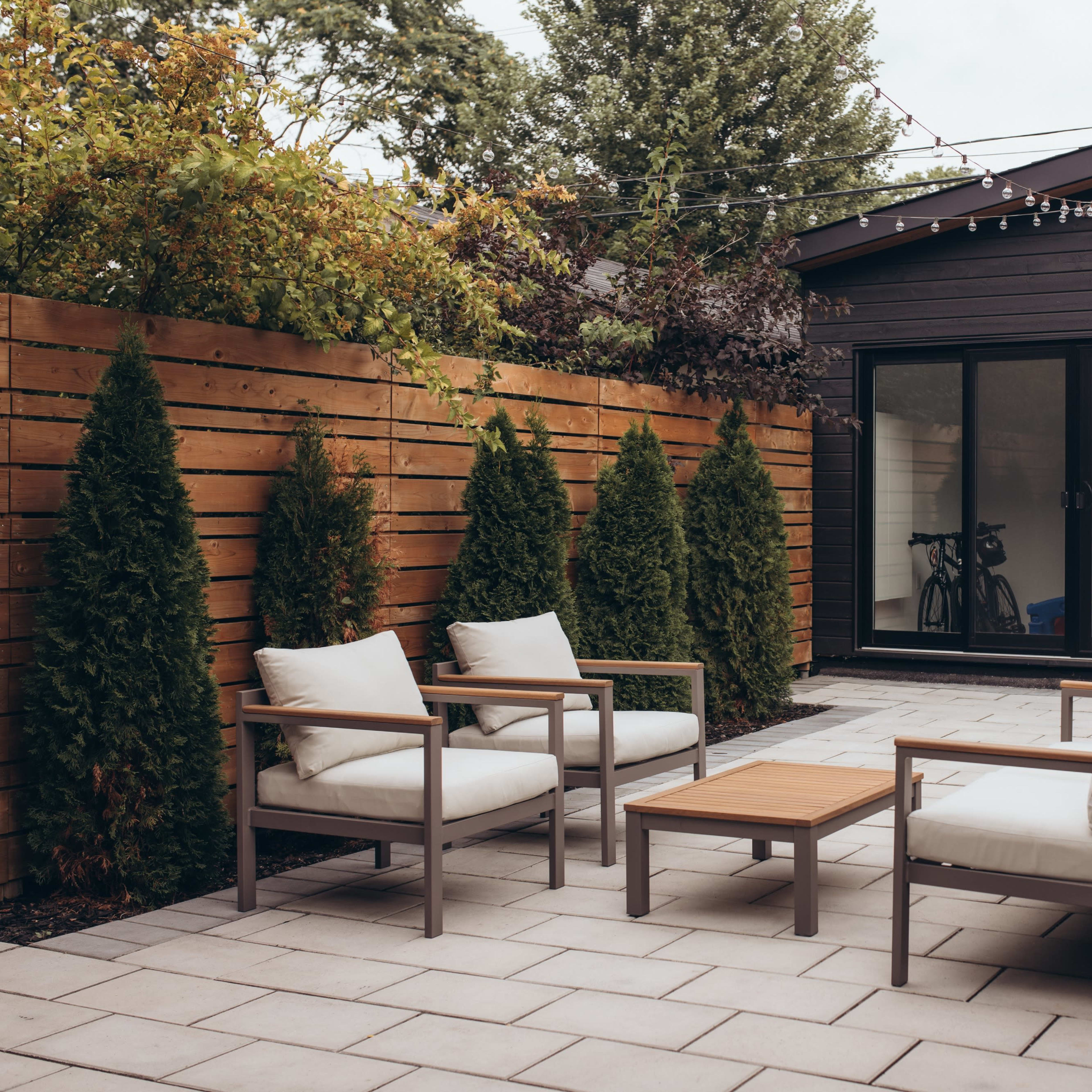A modern patio area with two chairs and a table surrounded by tall hedges and a building in the background.