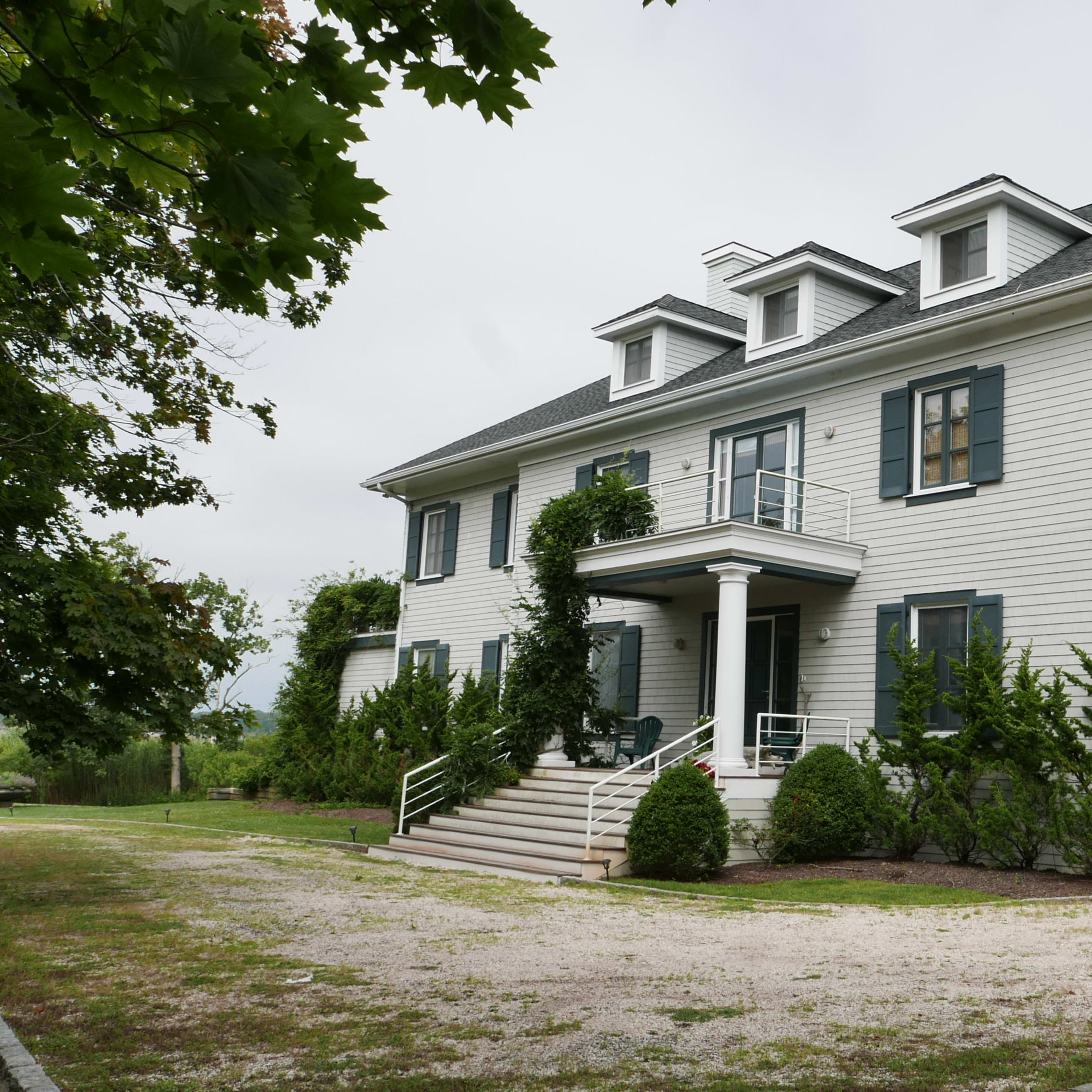 A large, two-story white house with multiple windows and a landscaped front yard sits along a gravel pathway.