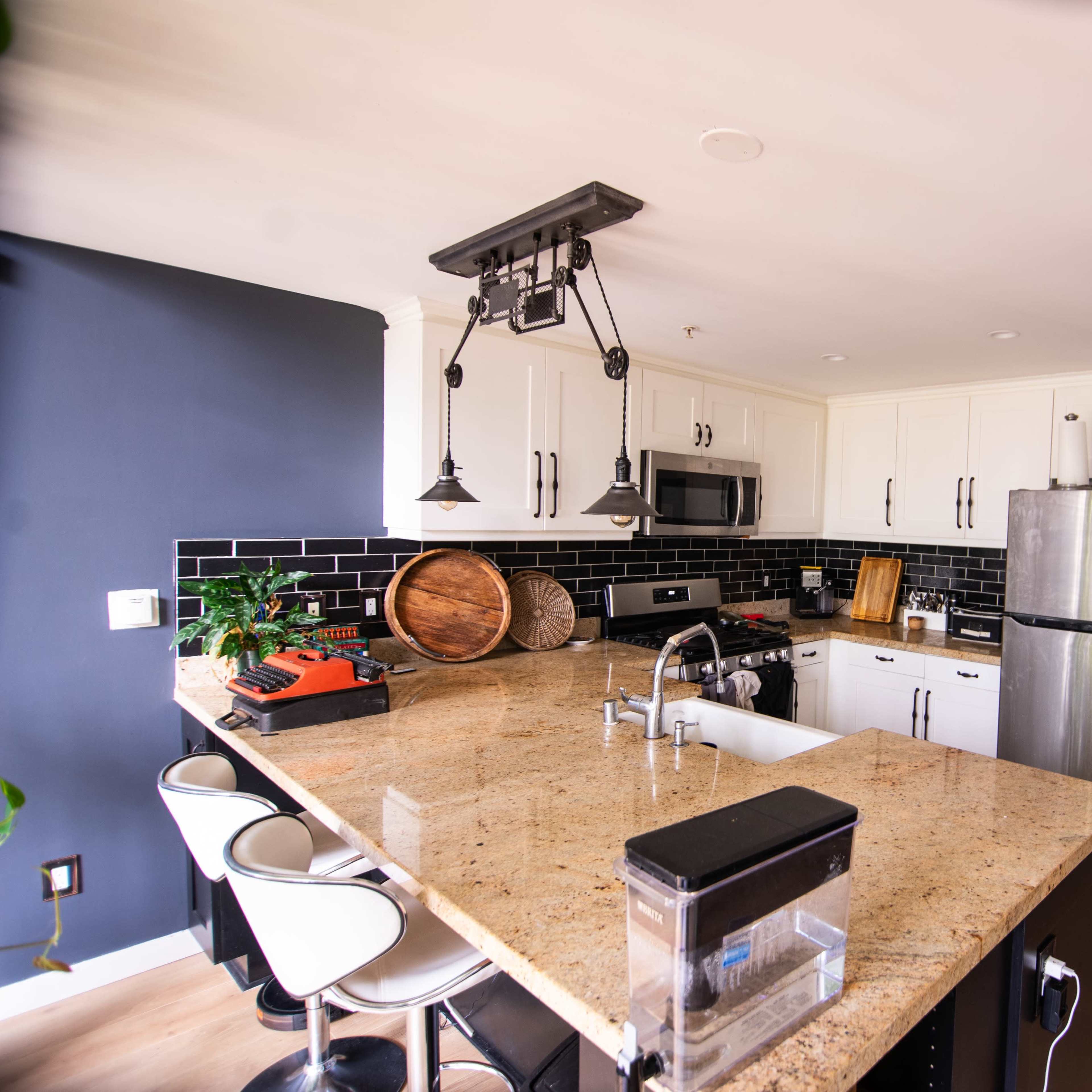 A modern kitchen features a large granite island, stainless steel appliances, and a black tile backsplash.