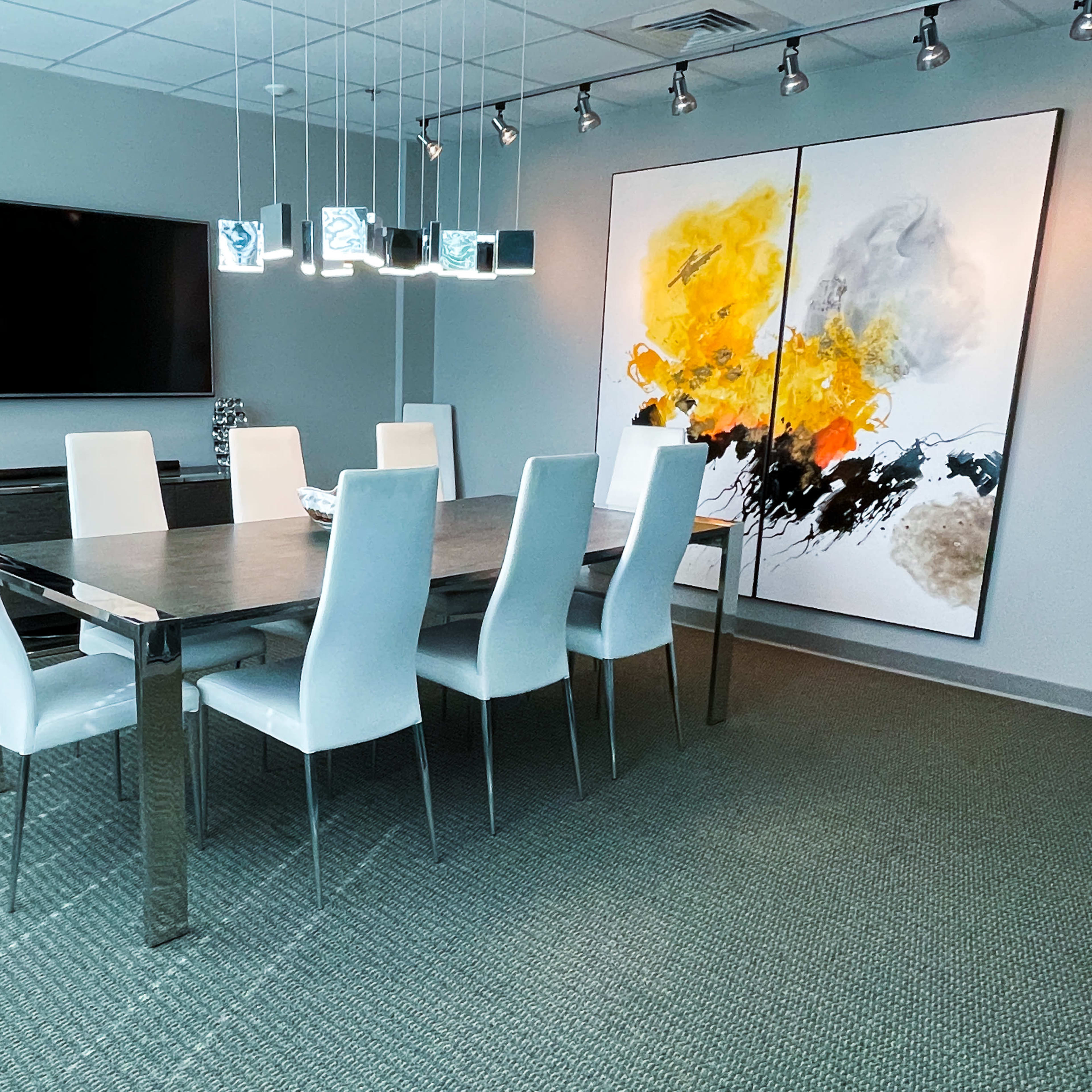 A modern conference room with a long table surrounded by white chairs, a large artwork on the wall, and a television mounted above a side cabinet.