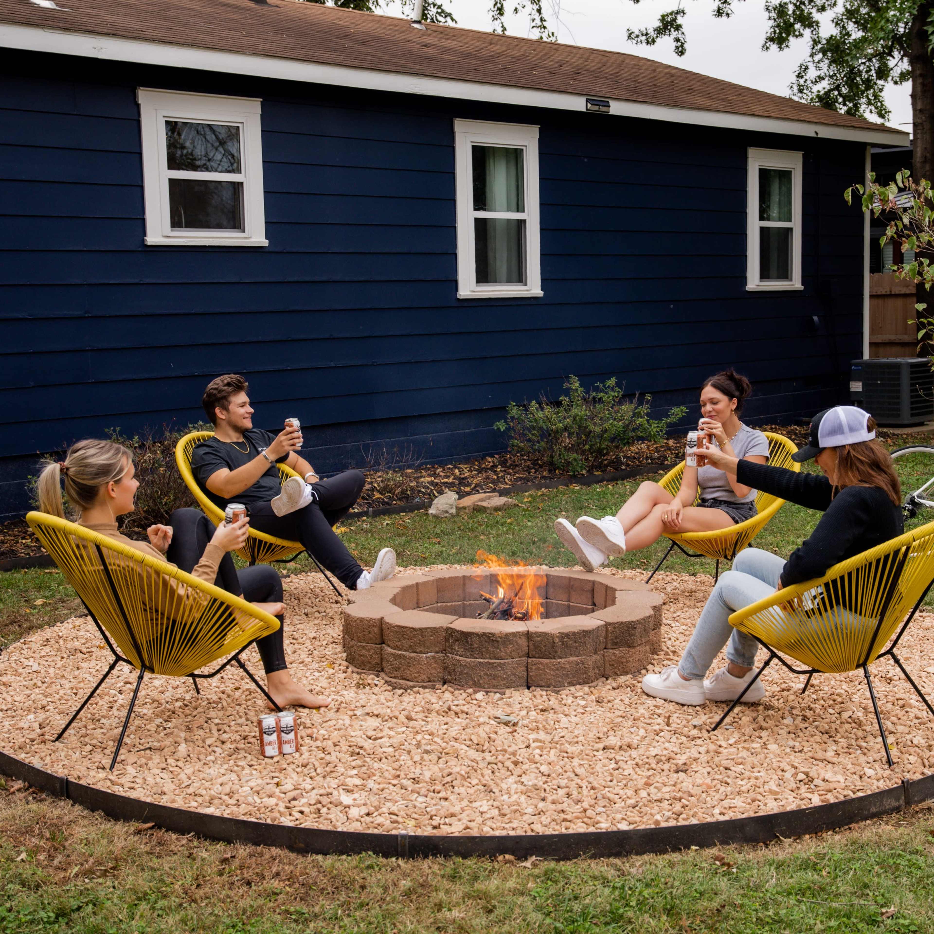 A group of four people sits around a fire pit on a gravel circle in a backyard, with a blue house and a bicycle visible in the background.