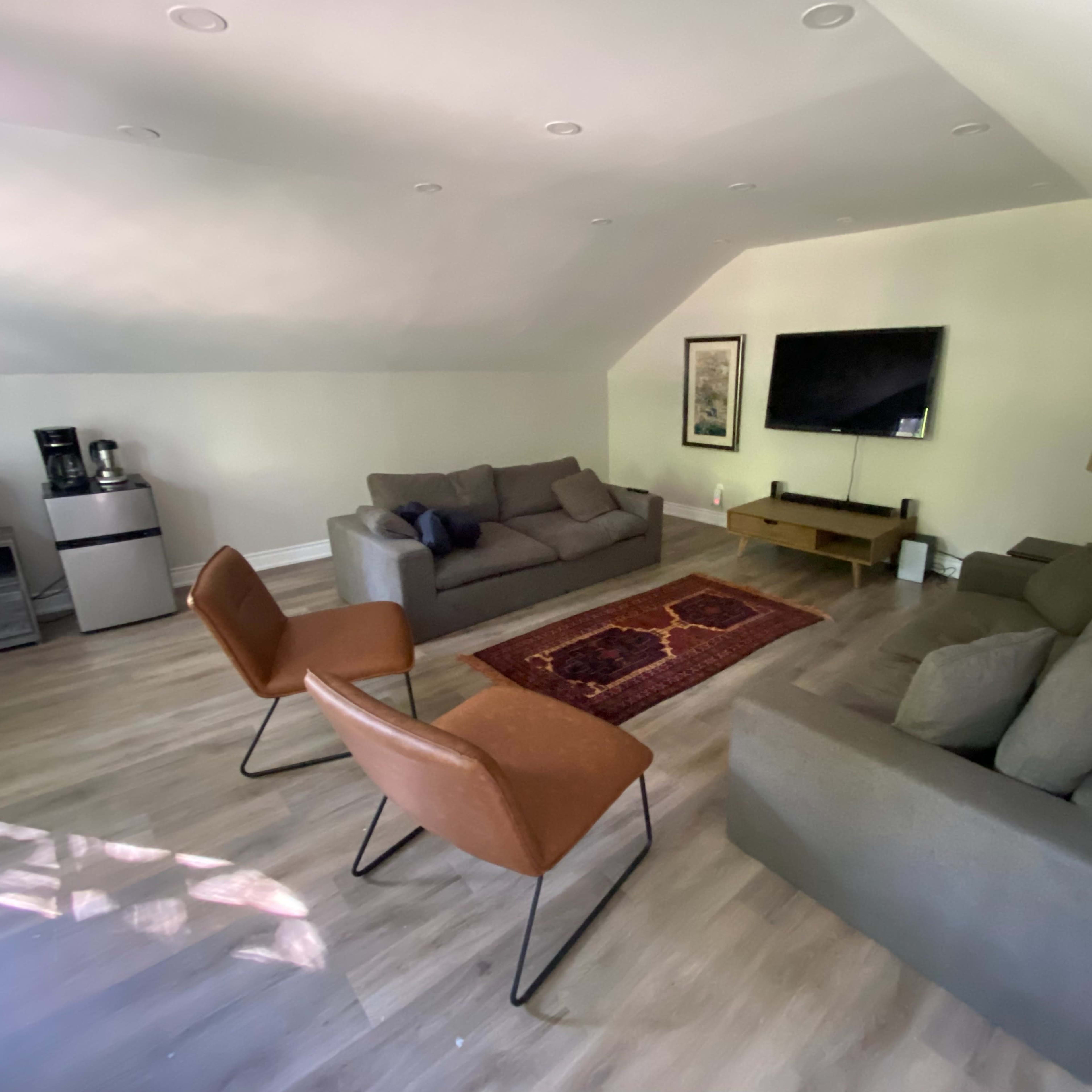 A spacious living area with two sofas, two chairs, a coffee table, a television mounted on the wall, and a kitchenette in the corner.