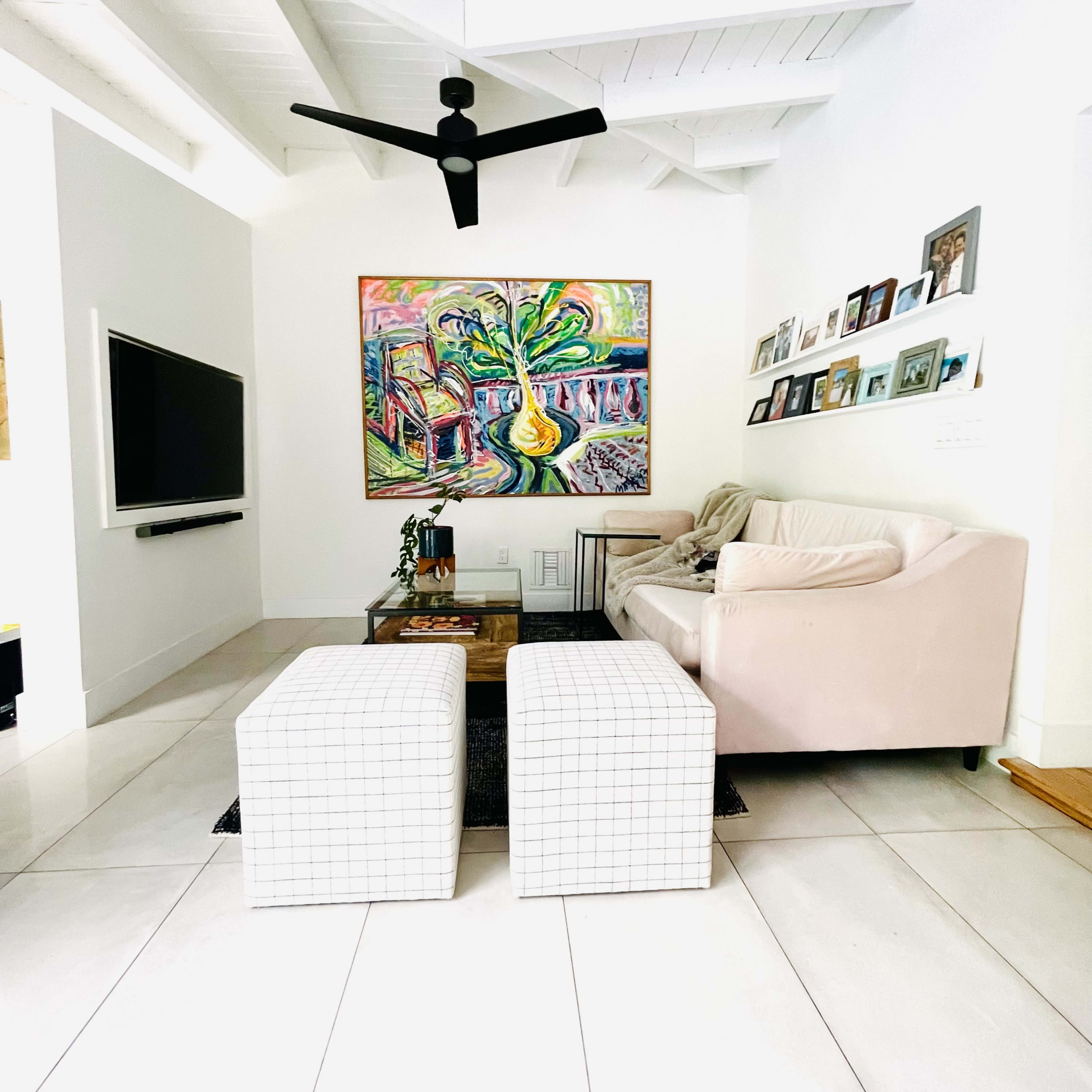 A modern living room features a pink couch, two square white ottomans, and a large colorful painting on the wall, with a television mounted opposite.