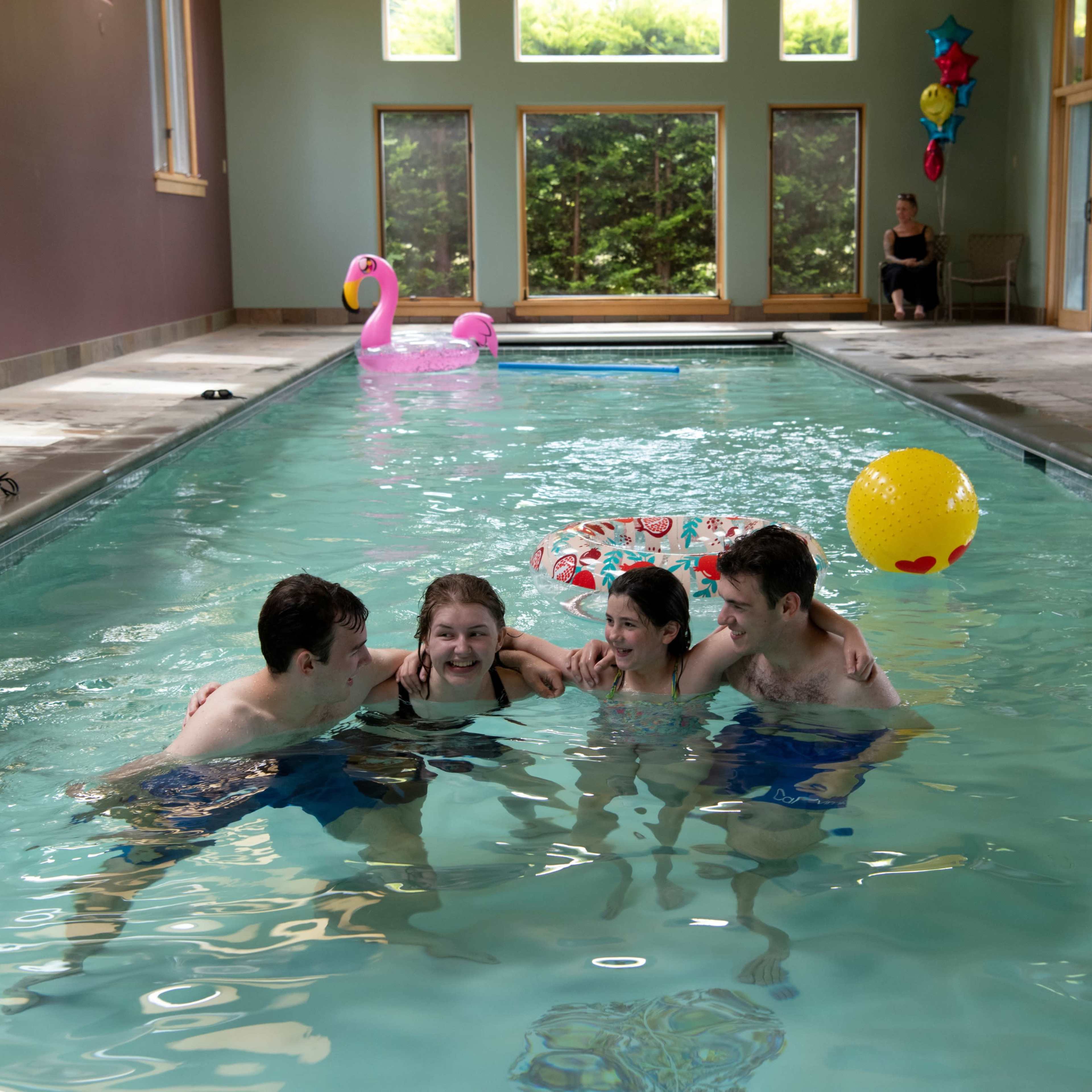 Four young people swim together in an indoor pool, while colorful decorations and floats are visible in the background.