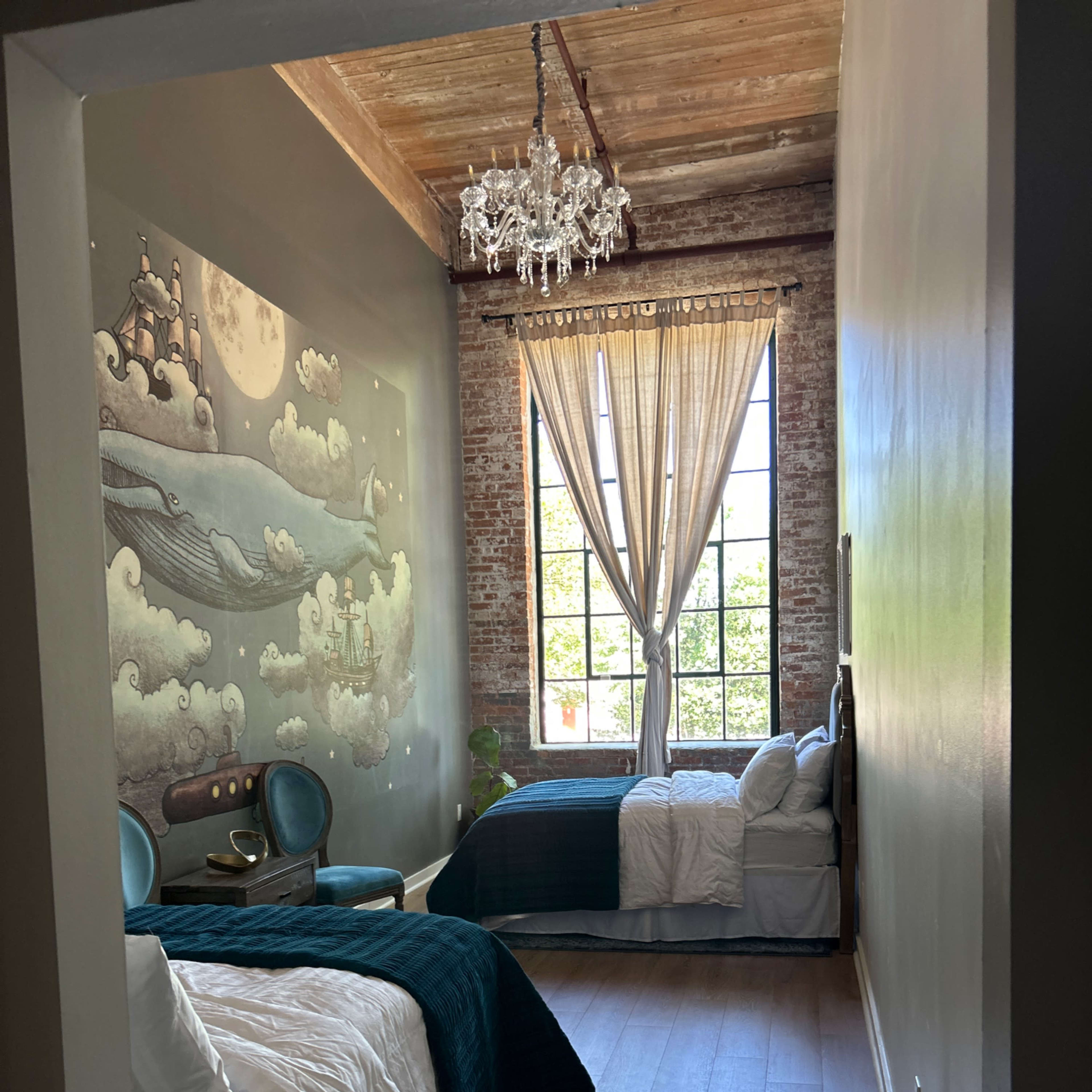 A spacious bedroom with two beds, a large window letting in natural light, and exposed brick walls decorated with a mural.