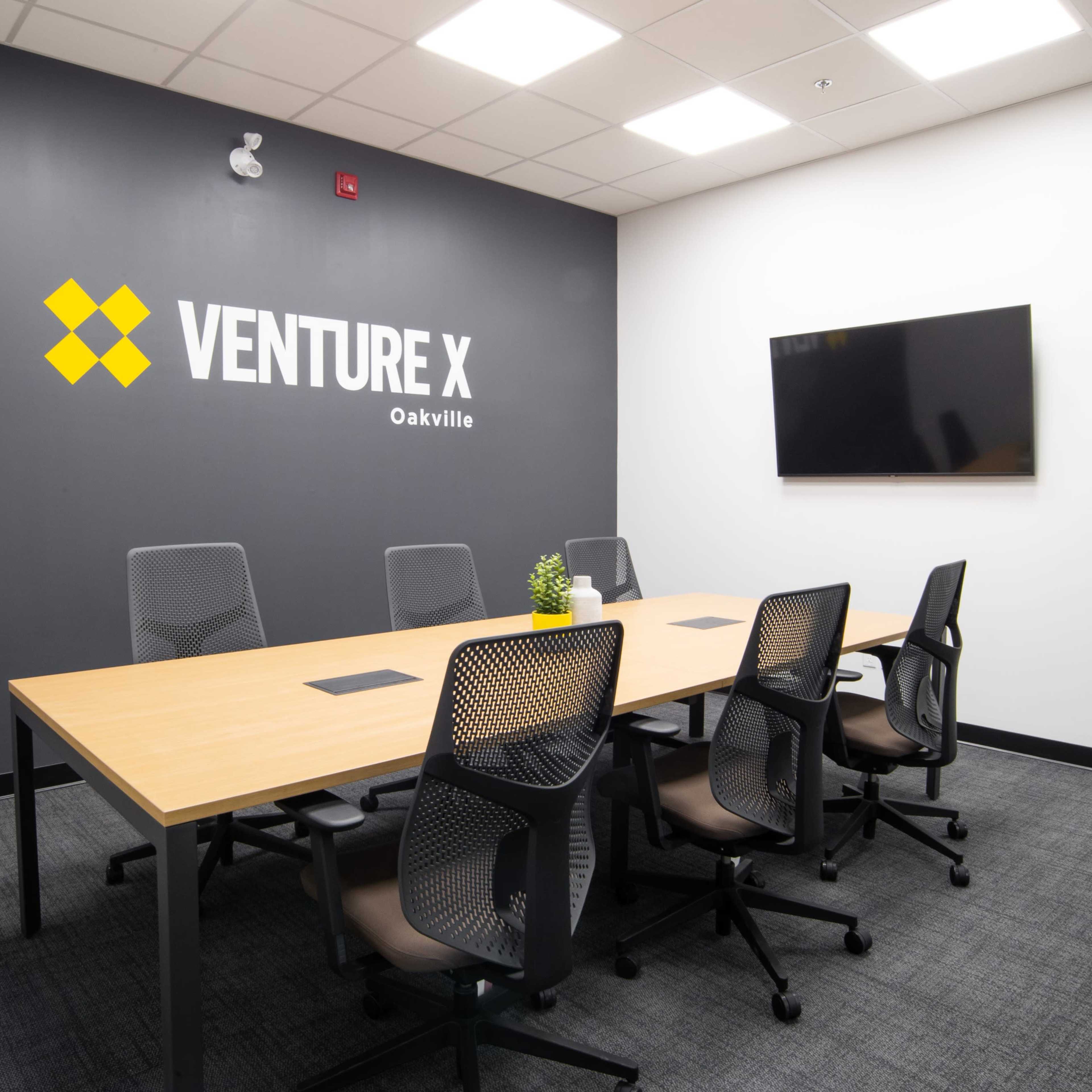 A modern conference room features a large wooden table surrounded by six ergonomic chairs, with a wall displaying the "VENTURE X Oakville" logo and a mounted television.