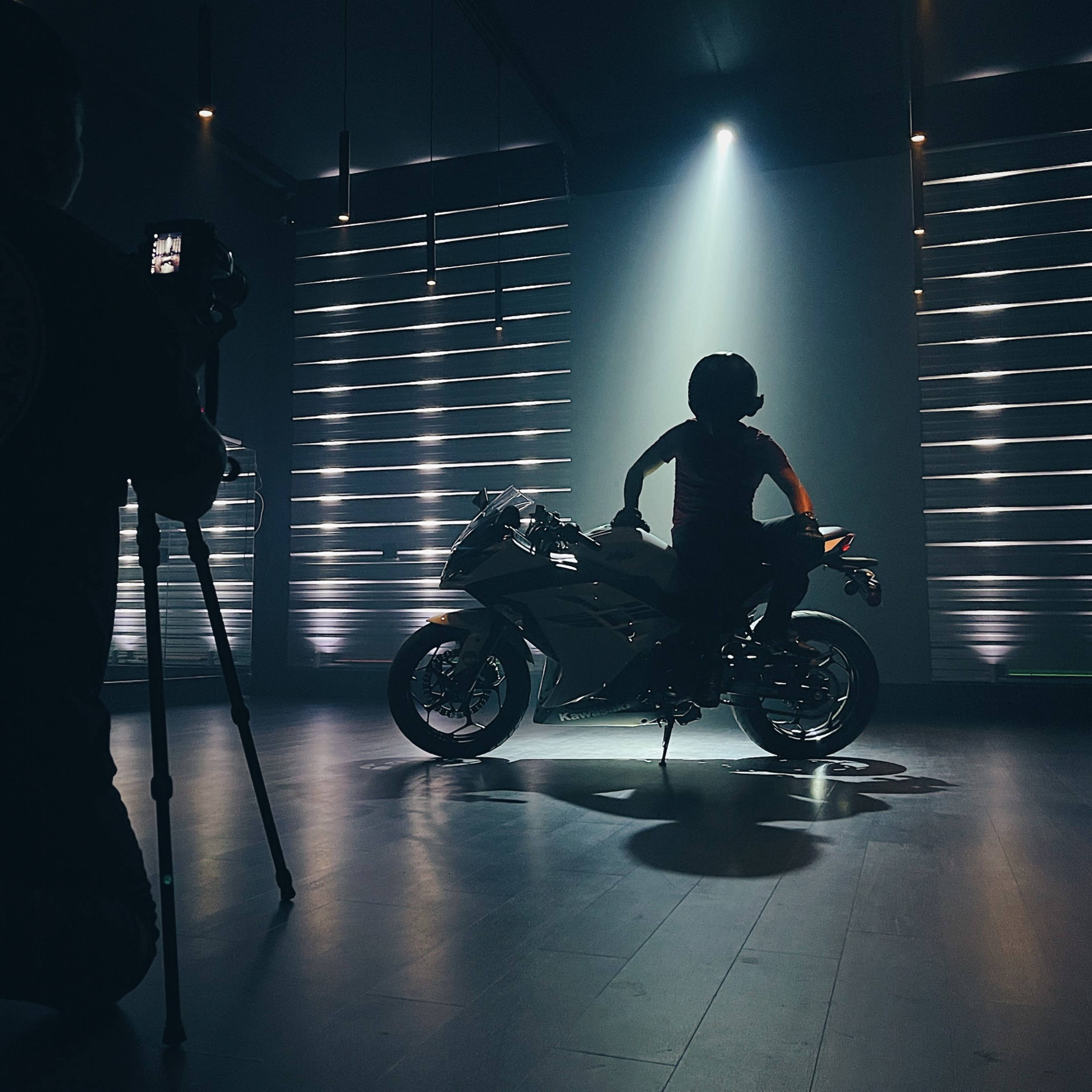 A person is photographing a motorcyclist posing on a motorcycle under dramatic stage lighting.