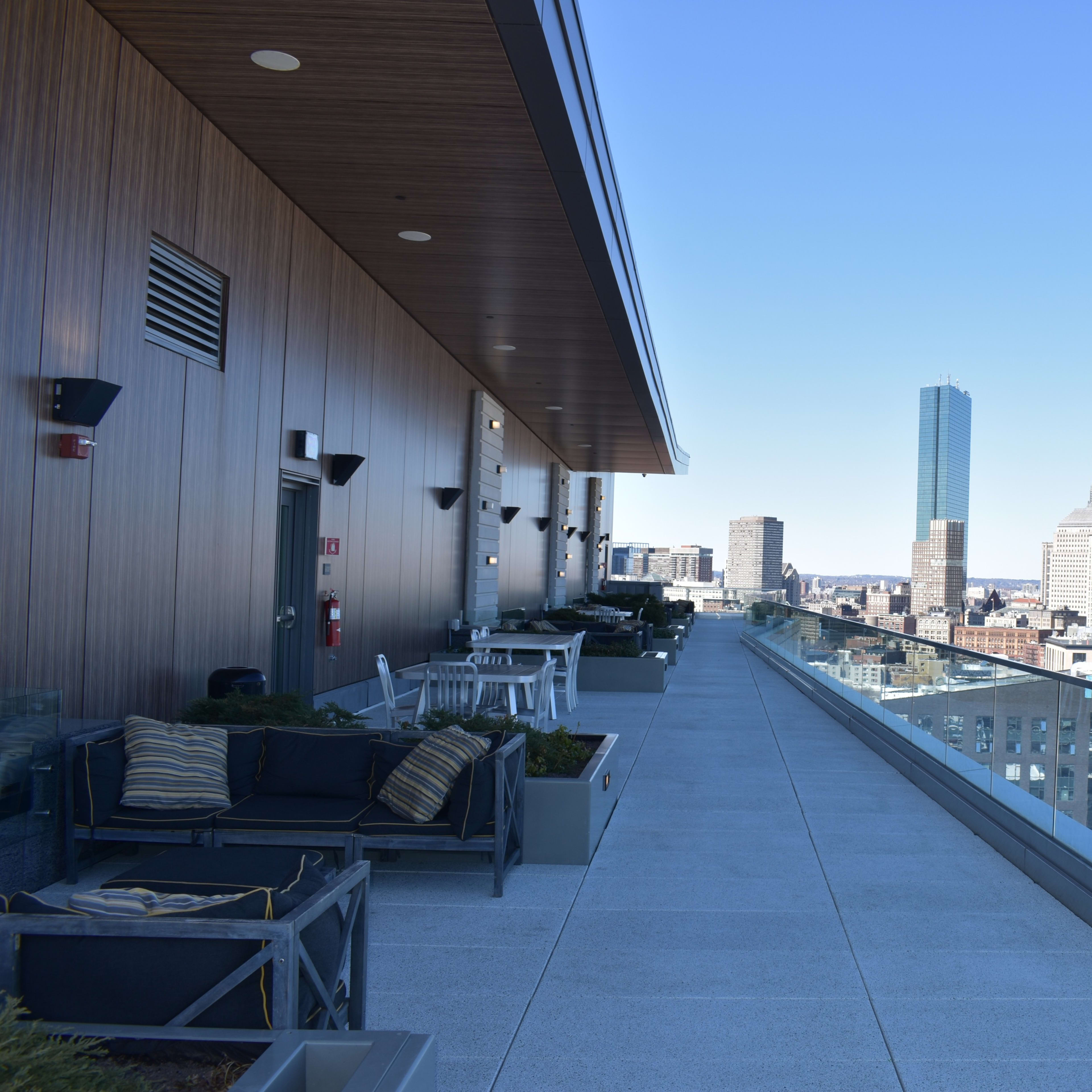 A spacious rooftop terrace with modern furniture, overlooking a city skyline featuring tall buildings and a clear blue sky.