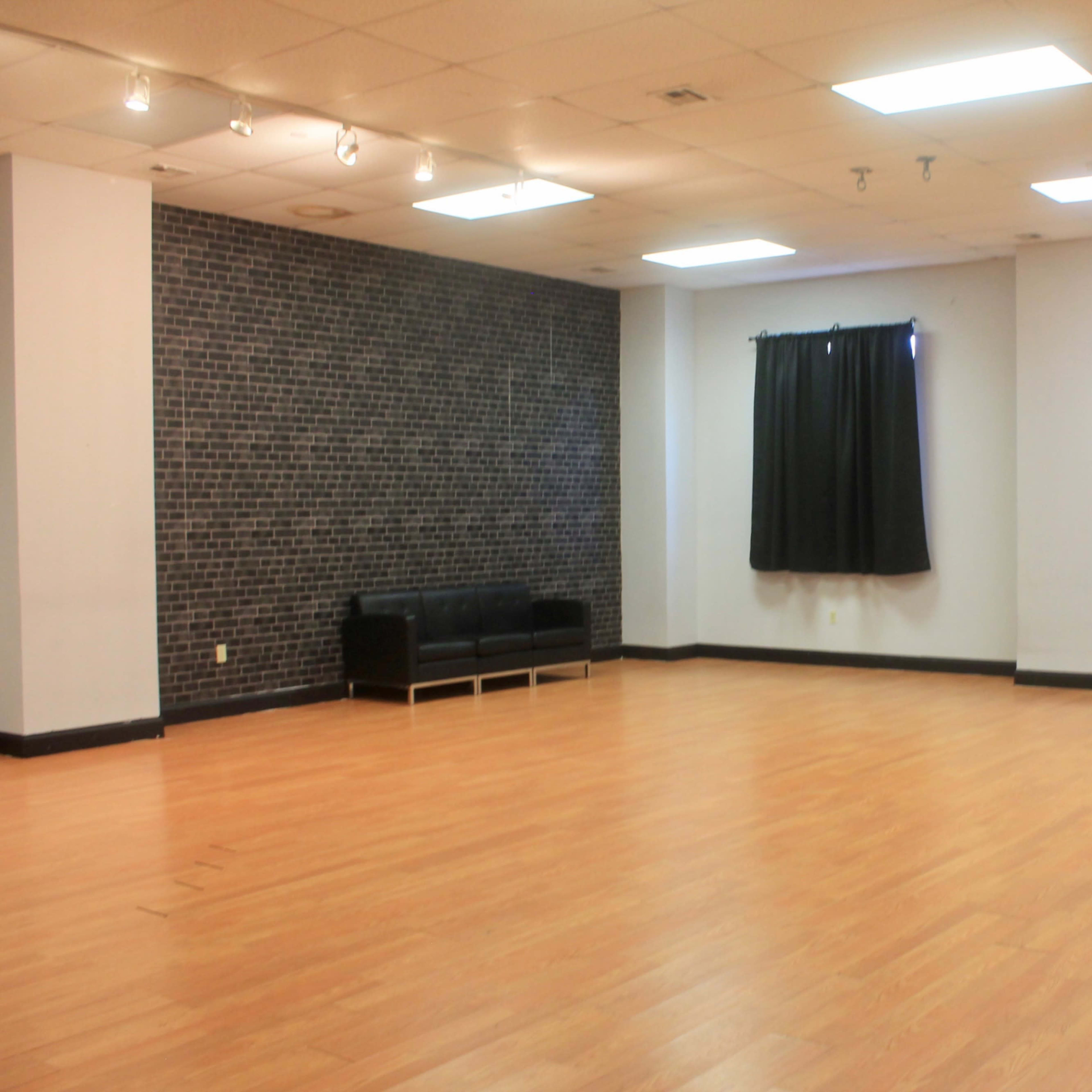 A spacious room with wooden flooring, a black sofa against a textured wall, a window covered by a black curtain, and a red exit door.