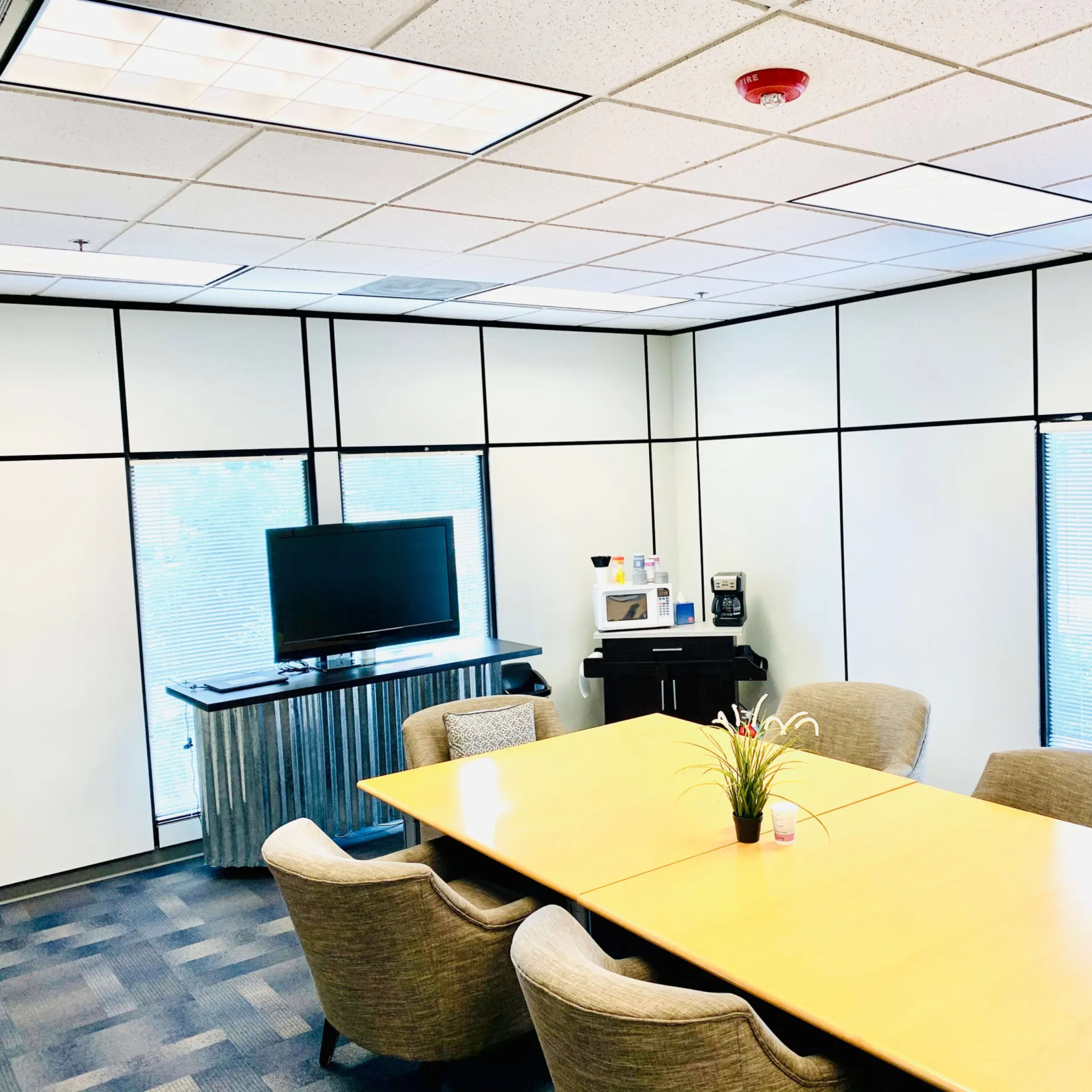 A conference room features a large wooden table surrounded by beige chairs, a television mounted on the wall, and a kitchenette area with a coffee machine.