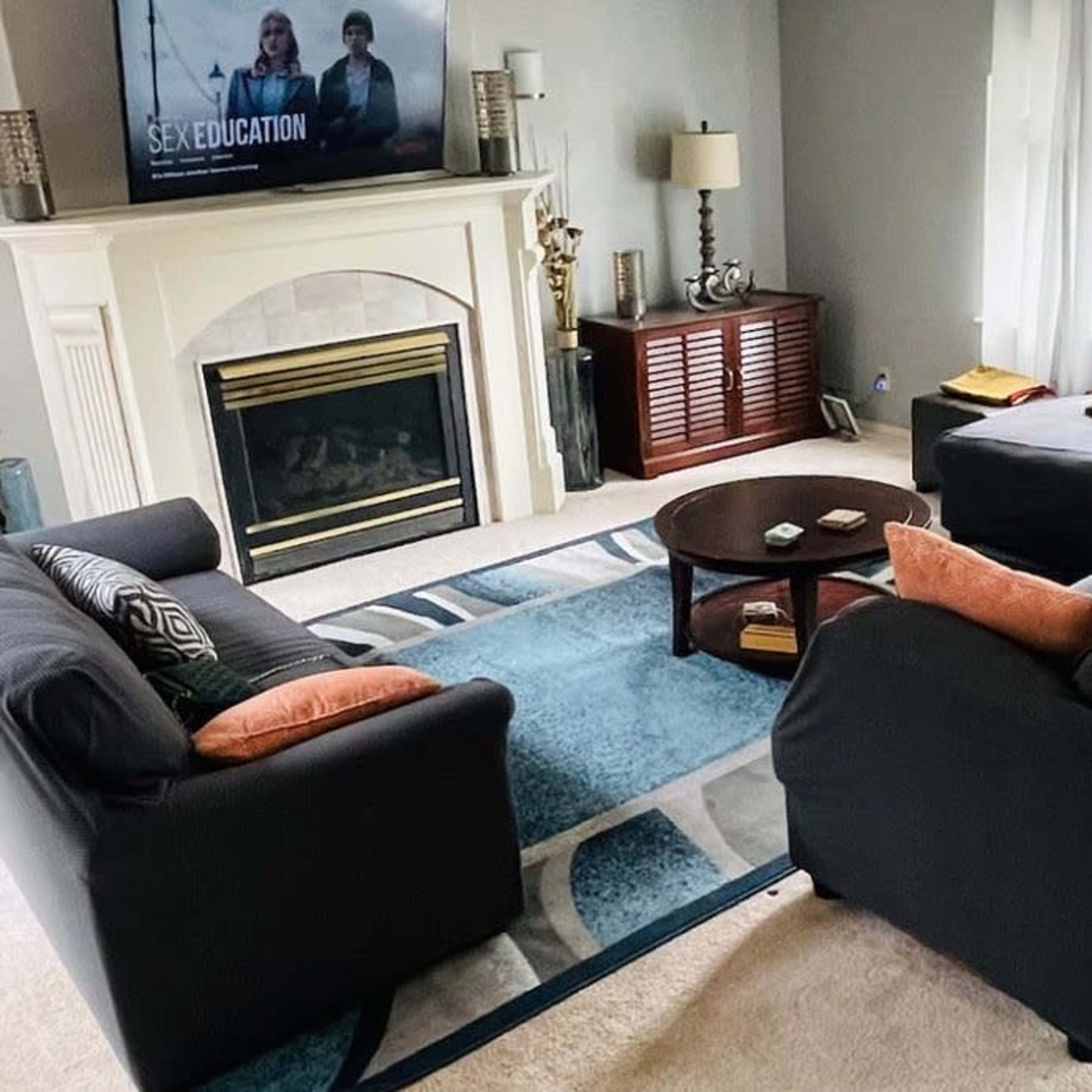The living room features two black couches with orange pillows facing a coffee table, a fireplace, and a television displaying the show "Sex Education."