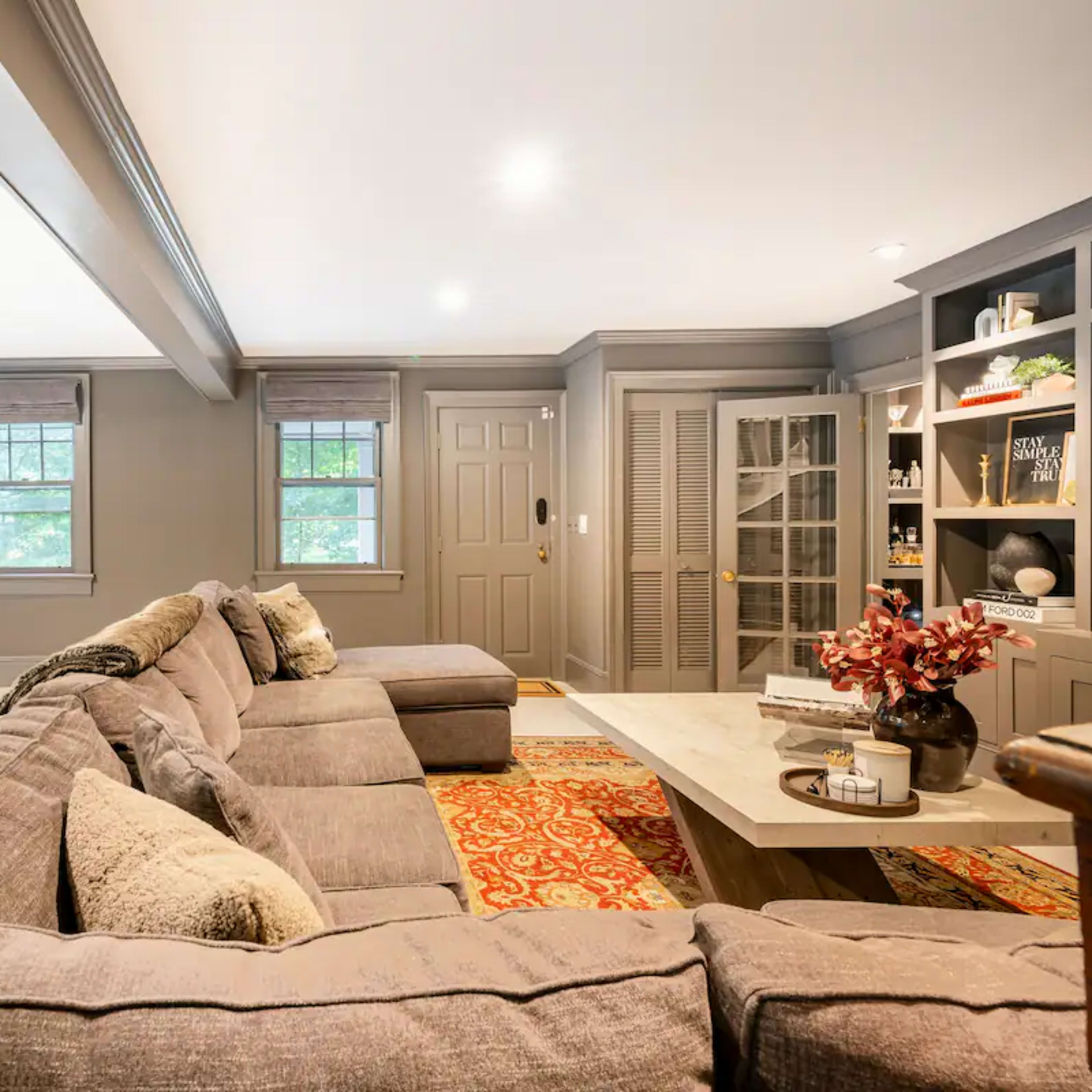 A modern living room features a large sectional sofa, a wooden dining table, and built-in shelves with decorative items.