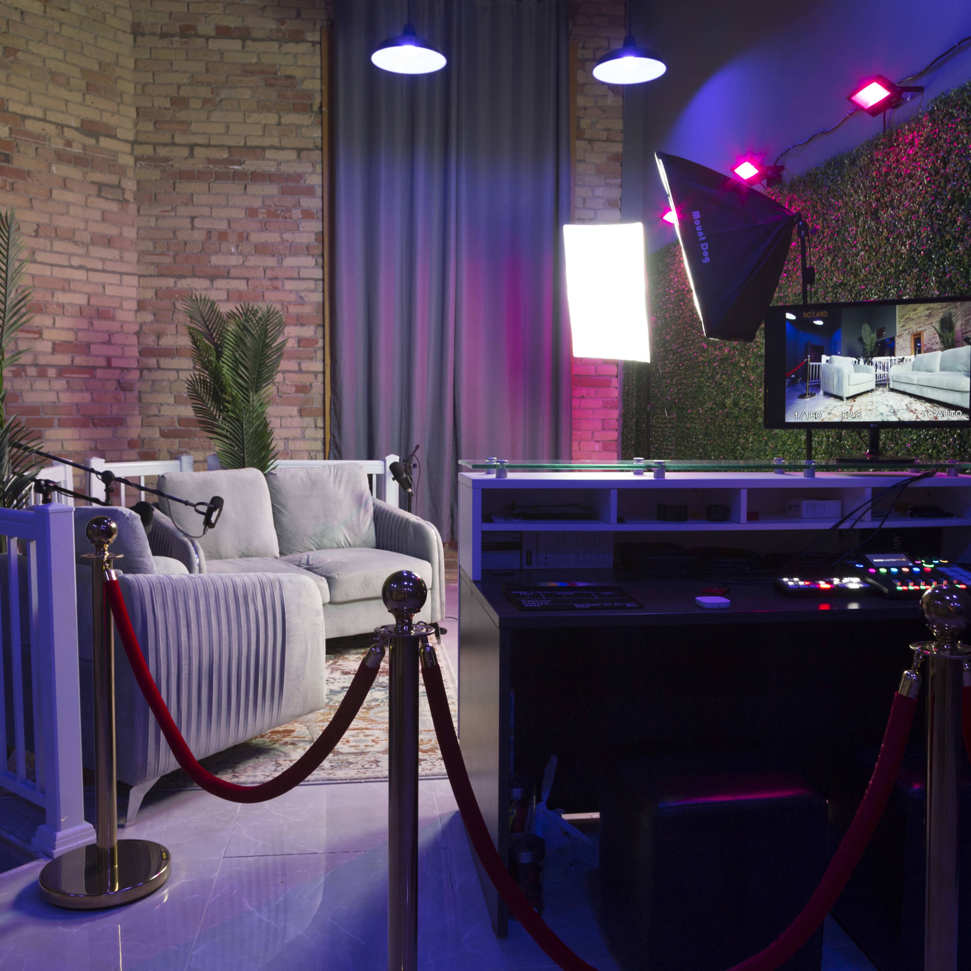 A media production studio featuring a cozy seating area with a backdrop of greenery and colorful lighting, along with a control desk and monitor.