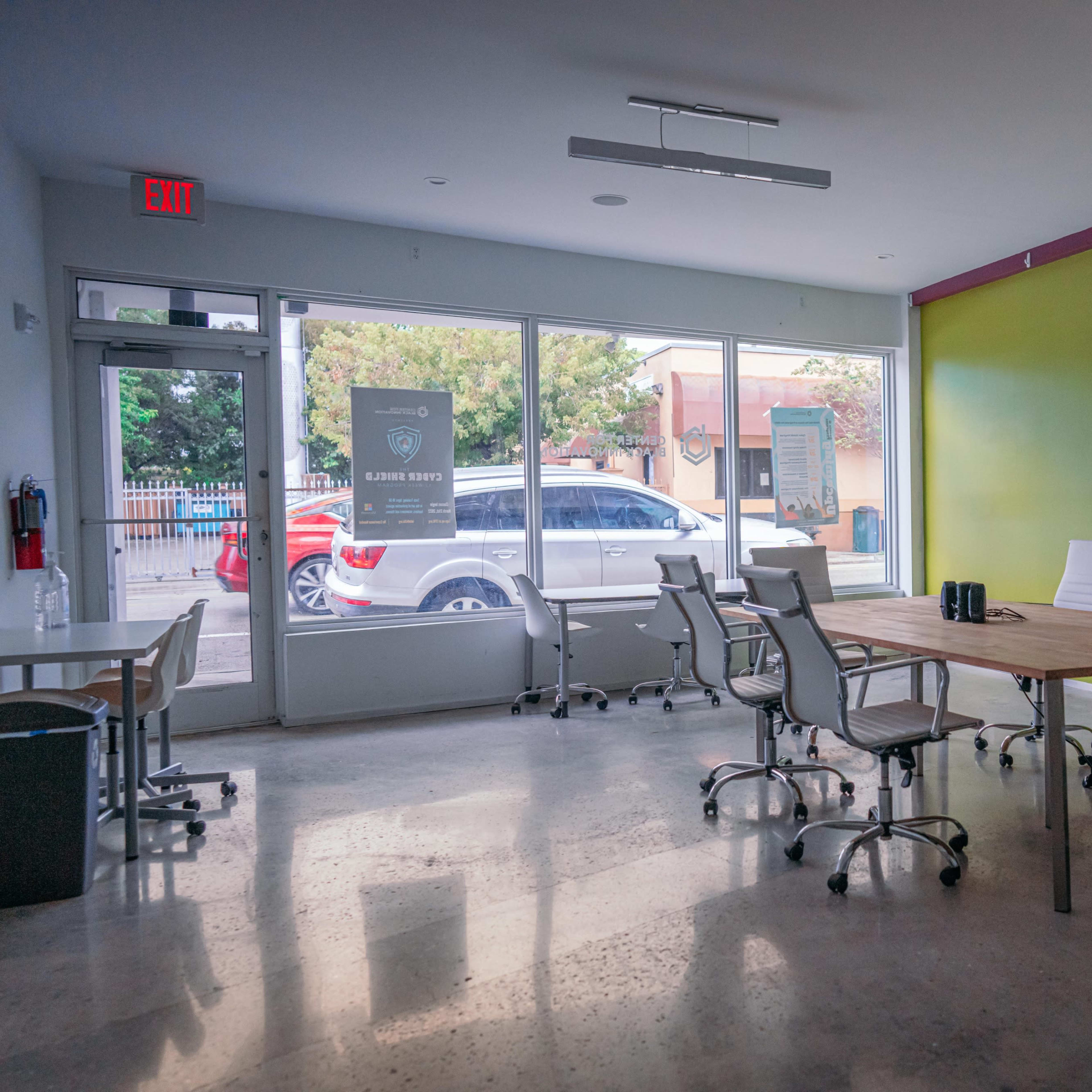 A modern conference room features a large wooden table surrounded by white chairs, with large windows offering a view of the street outside.