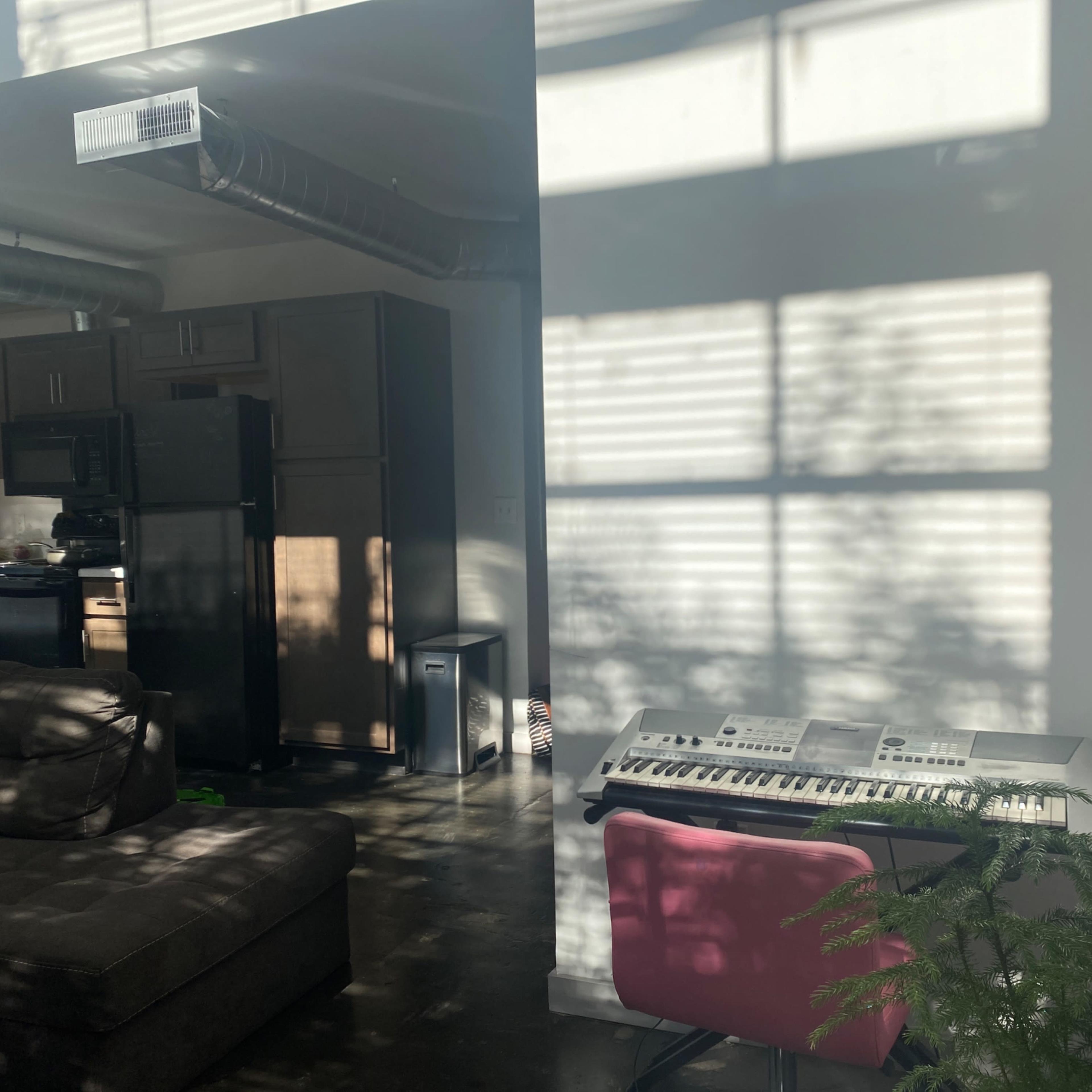 A keyboard sits next to a sofa in a living area illuminated by sunlight filtering through window blinds.