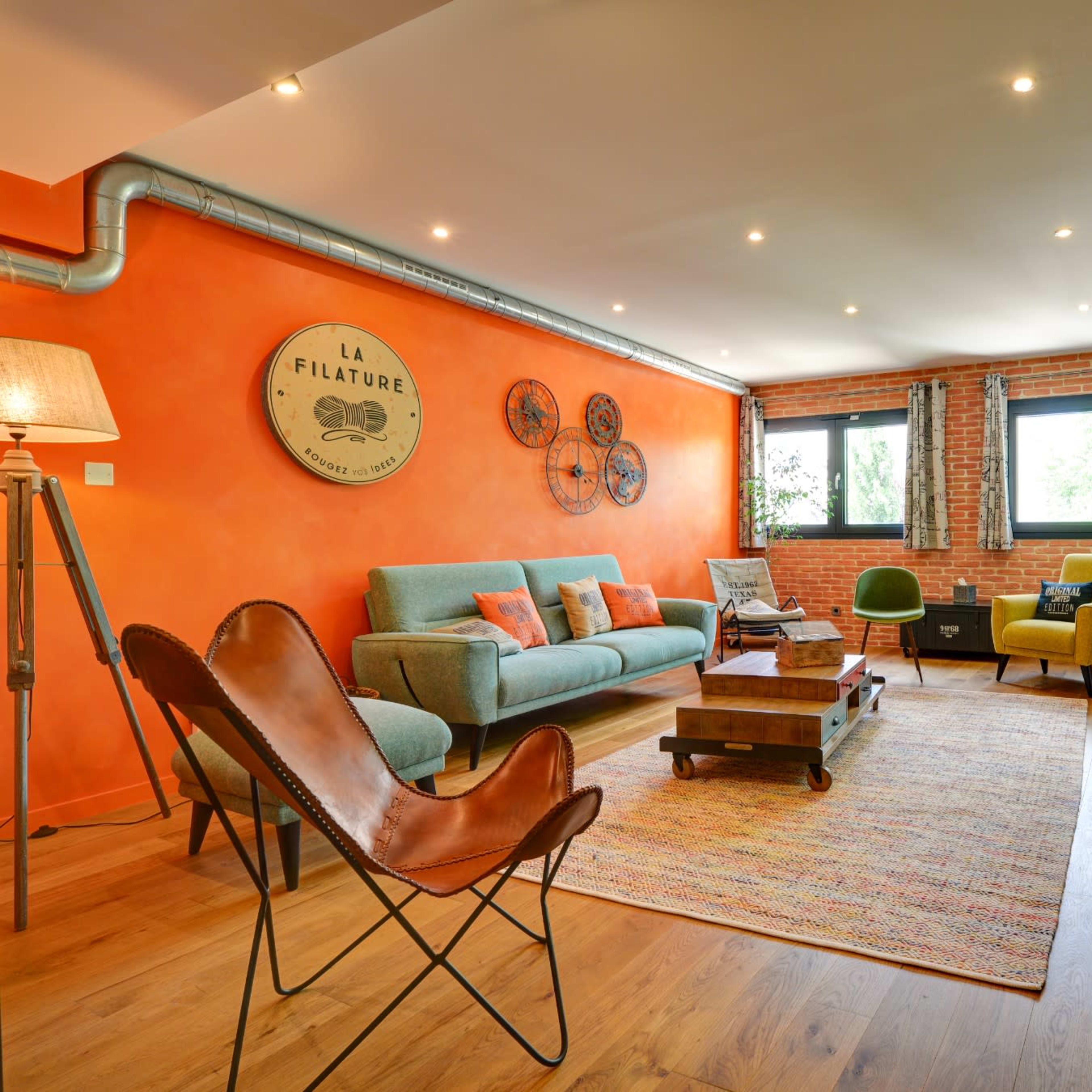 The room features a mix of seating options arranged on a colorful rug, with an orange accent wall and large windows providing natural light.