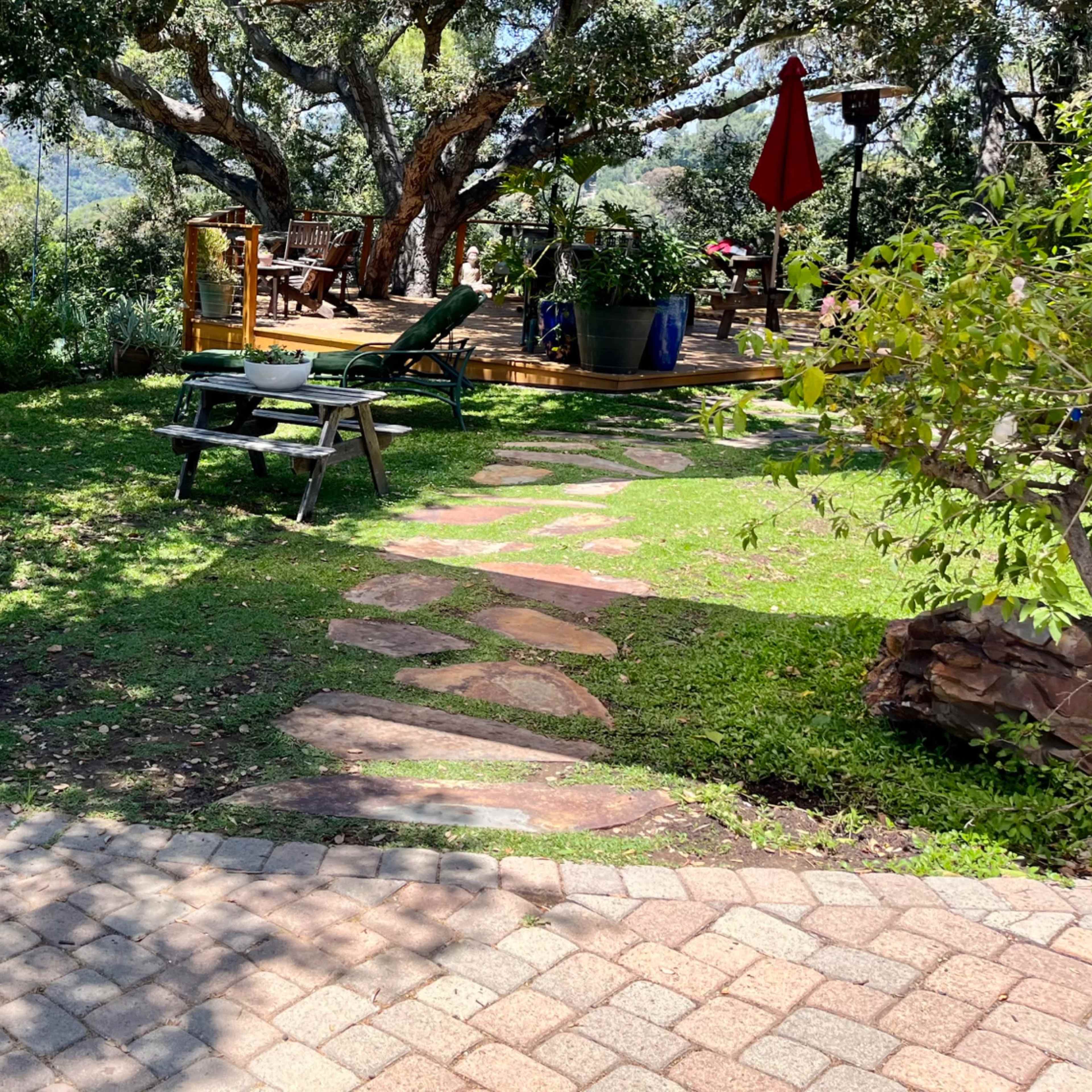 A stone pathway leads through a garden with lush greenery to a wooden deck shaded by large trees.