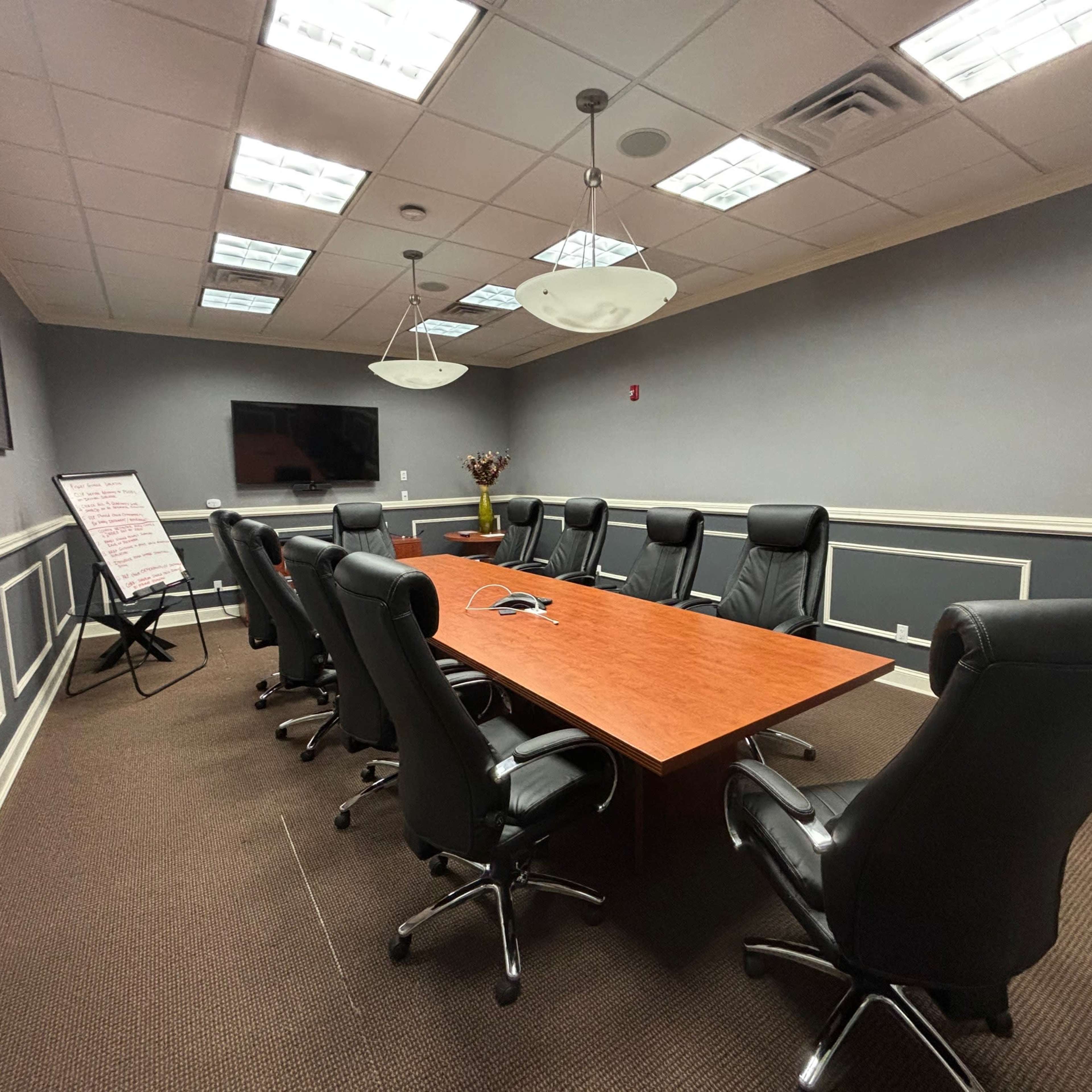 A conference room is furnished with a long wooden table surrounded by leather chairs, with a television on the wall and a flip chart nearby.