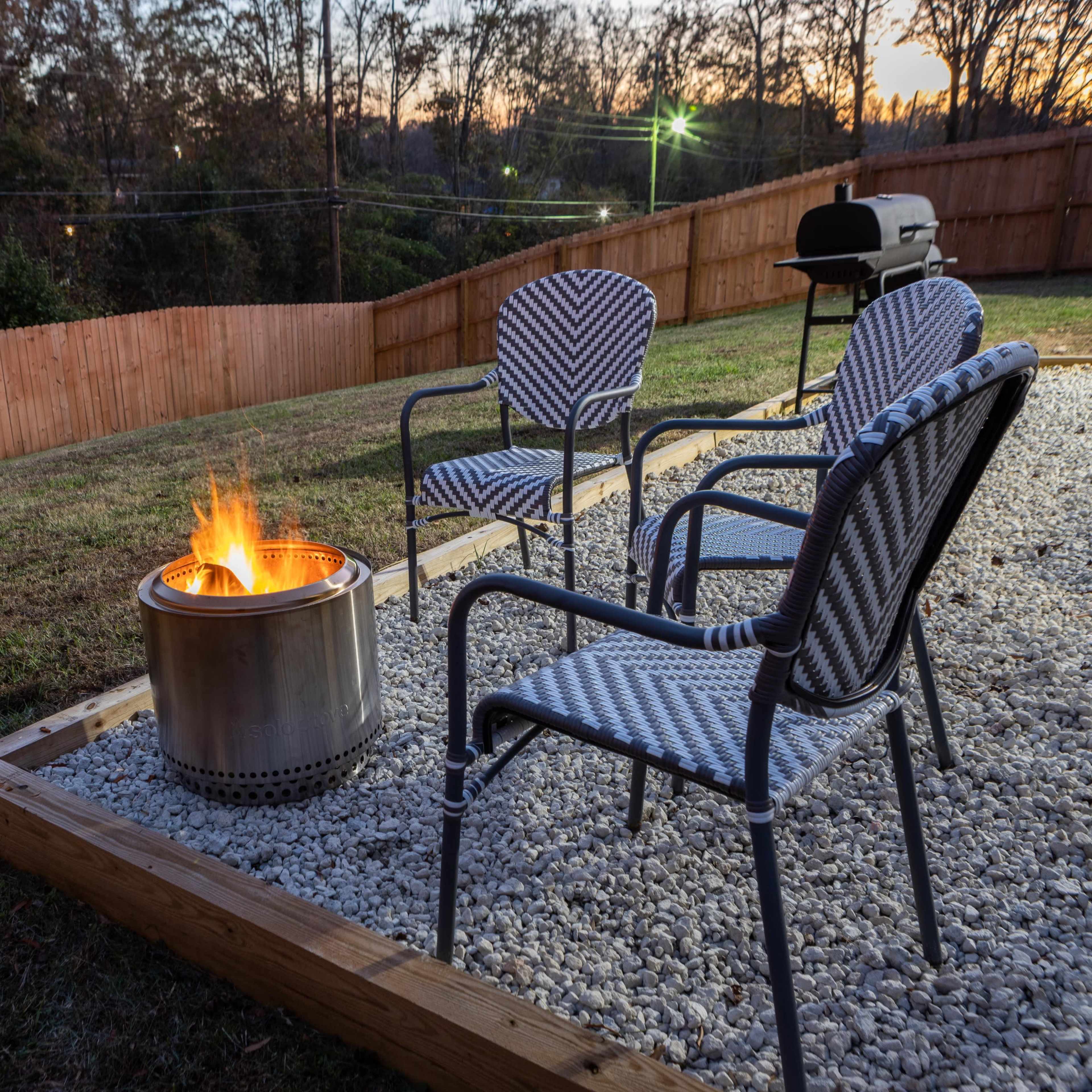 A backyard features a circular fire pit with flames, surrounded by three chairs, and a grill set against a wooden fence at dusk.