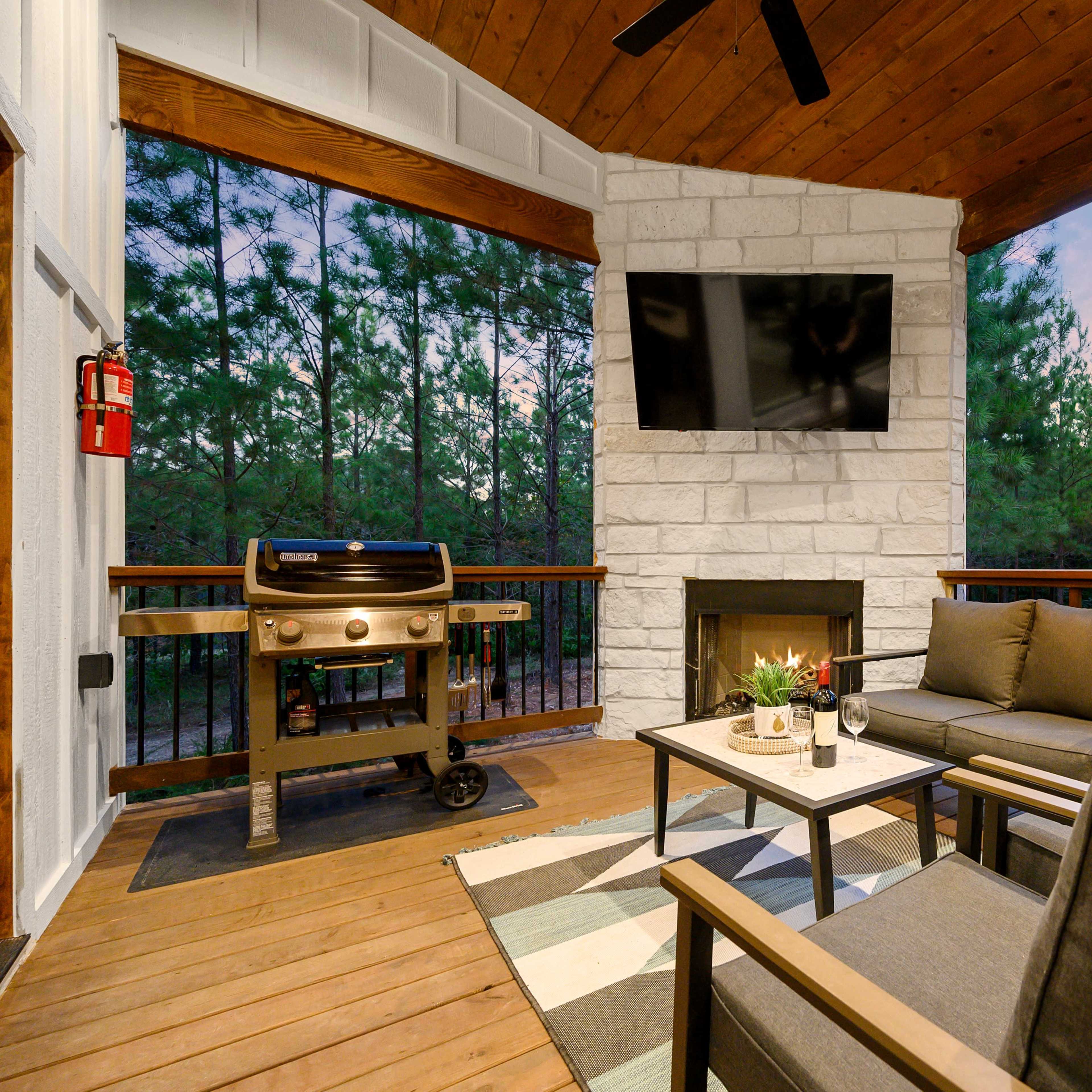 A covered outdoor living space featuring a grill, seating area with two chairs and a table, a stone fireplace, and a mounted television, all surrounded by wooded scenery.